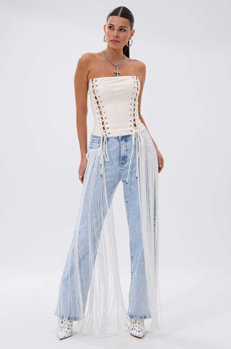 A woman poses in FLYING AWAY FRINGE DENIM, featuring white tassel details, paired with a strapless white lace-up corset top, silver necklace, and white boots against a plain white background.