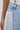 Close-up of a person wearing the FLYING AWAY FRINGE DENIM: light blue, high-rise jeans with white fringe details on the front pocket and a white lace-up corset belt. The person's hand rests relaxed by their side.