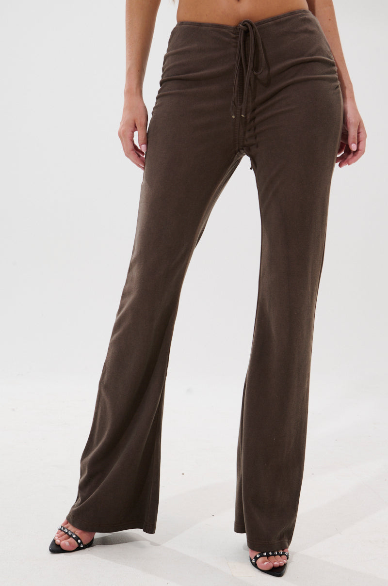 A person wears FESTIVAL LOW RISE RUCHED FLARE LEGGING in brown with a drawstring waist, styled with open-toed black heels, standing against a plain white background.