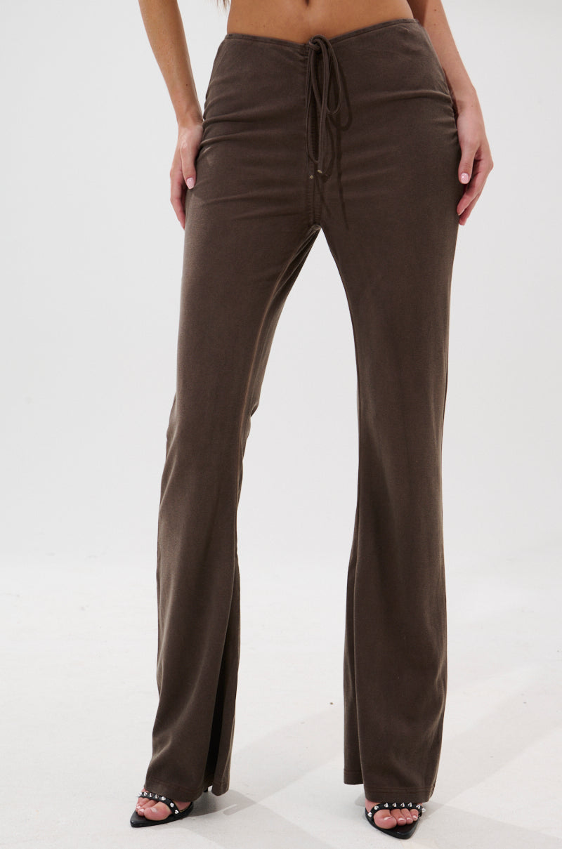 A person wears FESTIVAL LOW RISE RUCHED FLARE LEGGING—velvet-like, low-rise with a ruched front and ankle slits—paired with open-toe heels against a plain white background.