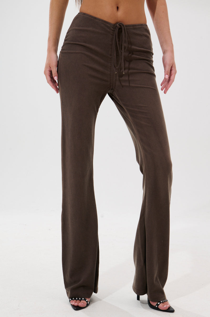 A person wearing brown FESTIVAL LOW RISE RUCHED FLARE LEGGING pants and black open-toe heels stands against a plain white background.