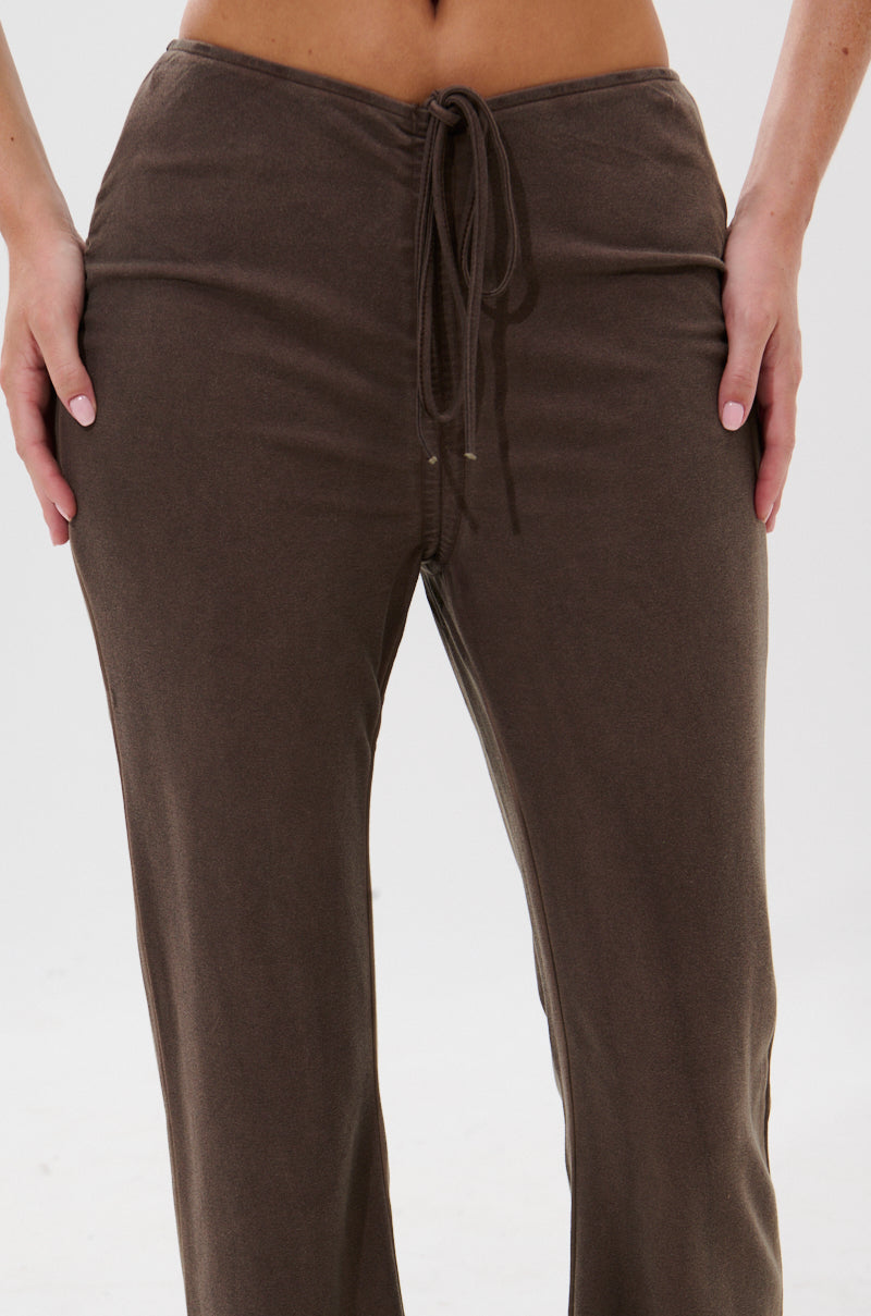 A close-up of a person wearing the FESTIVAL LOW RISE RUCHED FLARE LEGGING in brown, featuring a relaxed fit and flared design, standing against a plain light background with hands resting on their hips.