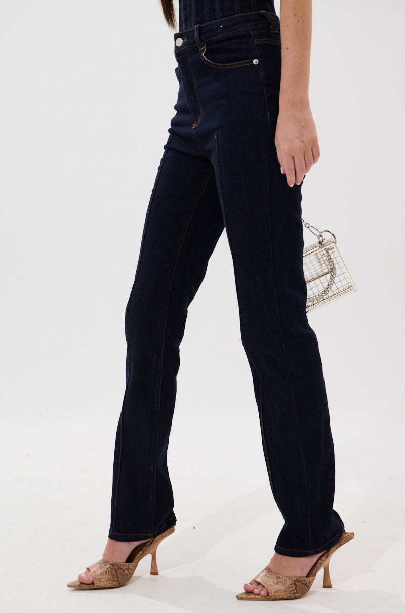 A person wearing GIVE IT TO ME BABY SKINNY DENIM in dark blue, beige heeled sandals, and holding a metallic handbag stands against a plain white background. 2