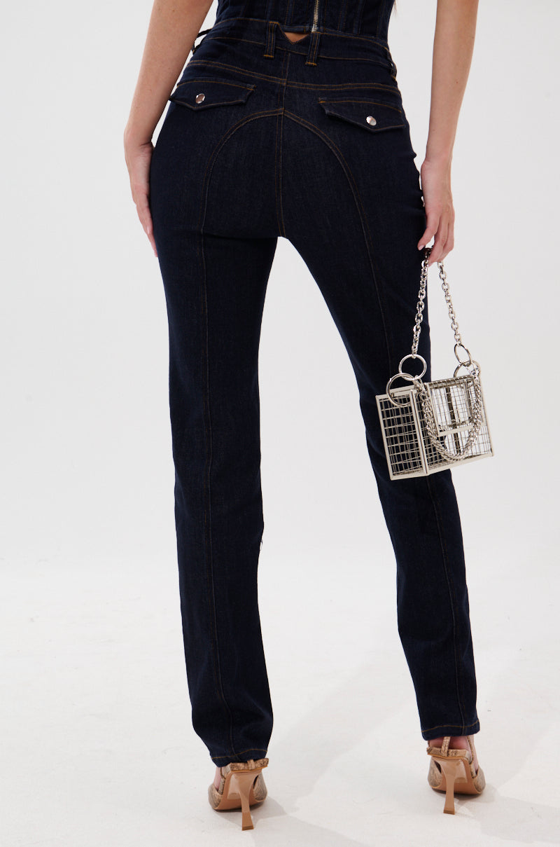A person stands facing away against a plain white background, wearing GIVE IT TO ME BABY SKINNY DENIM jeans and nude heels, holding a small silver cage-shaped purse by their side. 3