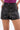 Wearing the SET IT OFF FAUX LEATHER SHORT IN BLACK with front pockets and a silver button, paired with a purple top. Only the lower torso and upper legs in these high-waisted, structured black shorts are visible.