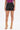 Wearing the SET IT OFF FAUX LEATHER SHORT IN BLACK, a person pairs them with a purple top and gold chain-strap high-heeled sandals, standing against a white background. Photo is cropped just below the shoulders.
