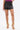 Model showcases the SET IT OFF FAUX LEATHER SHORT IN BLACK, paired with gold high-heeled sandals featuring chunky chain details. Only the lower half is visible against a plain white background.