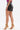 Wearing the SET IT OFF FAUX LEATHER SHORT IN BLACK, a purple sleeveless top, and gold high-heeled sandals with chunky chain details, a person poses against a plain white background.