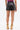 A woman faces away against a white background, wearing the SET IT OFF FAUX LEATHER SHORT IN BLACK, paired with a purple top and yellow high-heeled sandals featuring gold chain details.