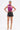A woman stands confidently in a shiny purple halter top, SET IT OFF FAUX LEATHER SHORT IN BLACK, gold strappy heels, and gold arm cuffs. Her hair is tied back as she poses against a plain white background.