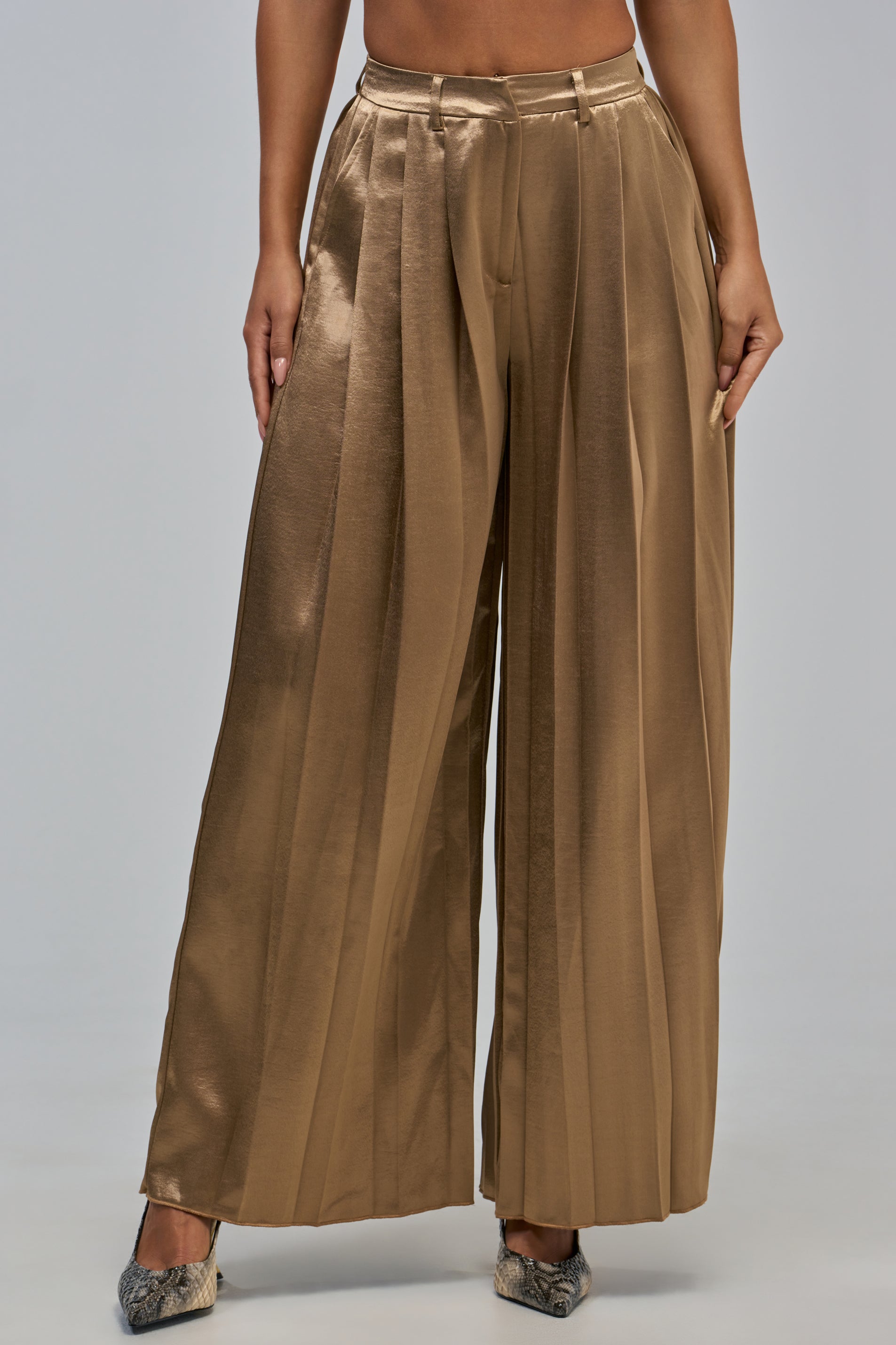 A person showcases chic evening style against a plain light gray background, wearing the IRIS SATIN PLEATED PANT IN LIGHT BROWN—wide-leg, high-waisted pants with pleats that elegantly catch the light.