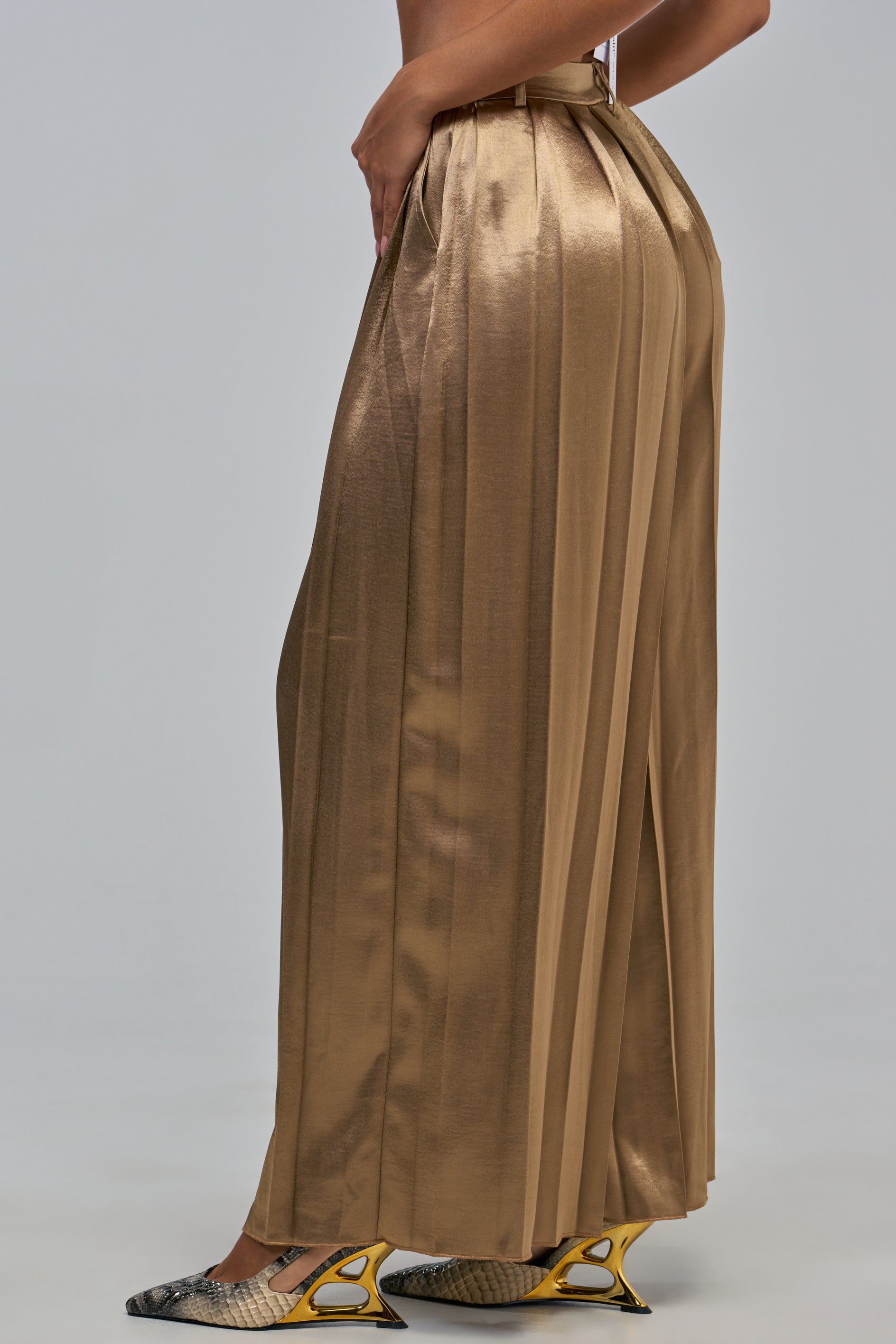 A person stands in profile, showcasing chic evening styling in the IRIS SATIN PLEATED PANT IN LIGHT BROWN with gray snake-pattern high-heeled shoes. The background is plain and light gray.
