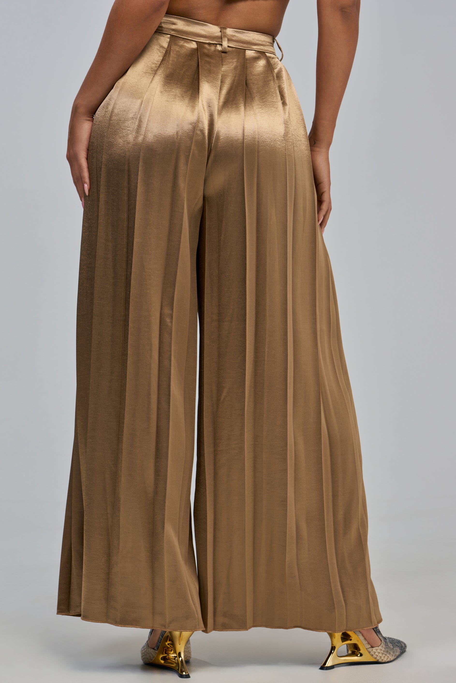 A person wearing the IRIS SATIN PLEATED PANT IN LIGHT BROWN, styled with metallic pointed heels and photographed from the back against a plain backdrop—an effortless look perfect for chic evening styling.