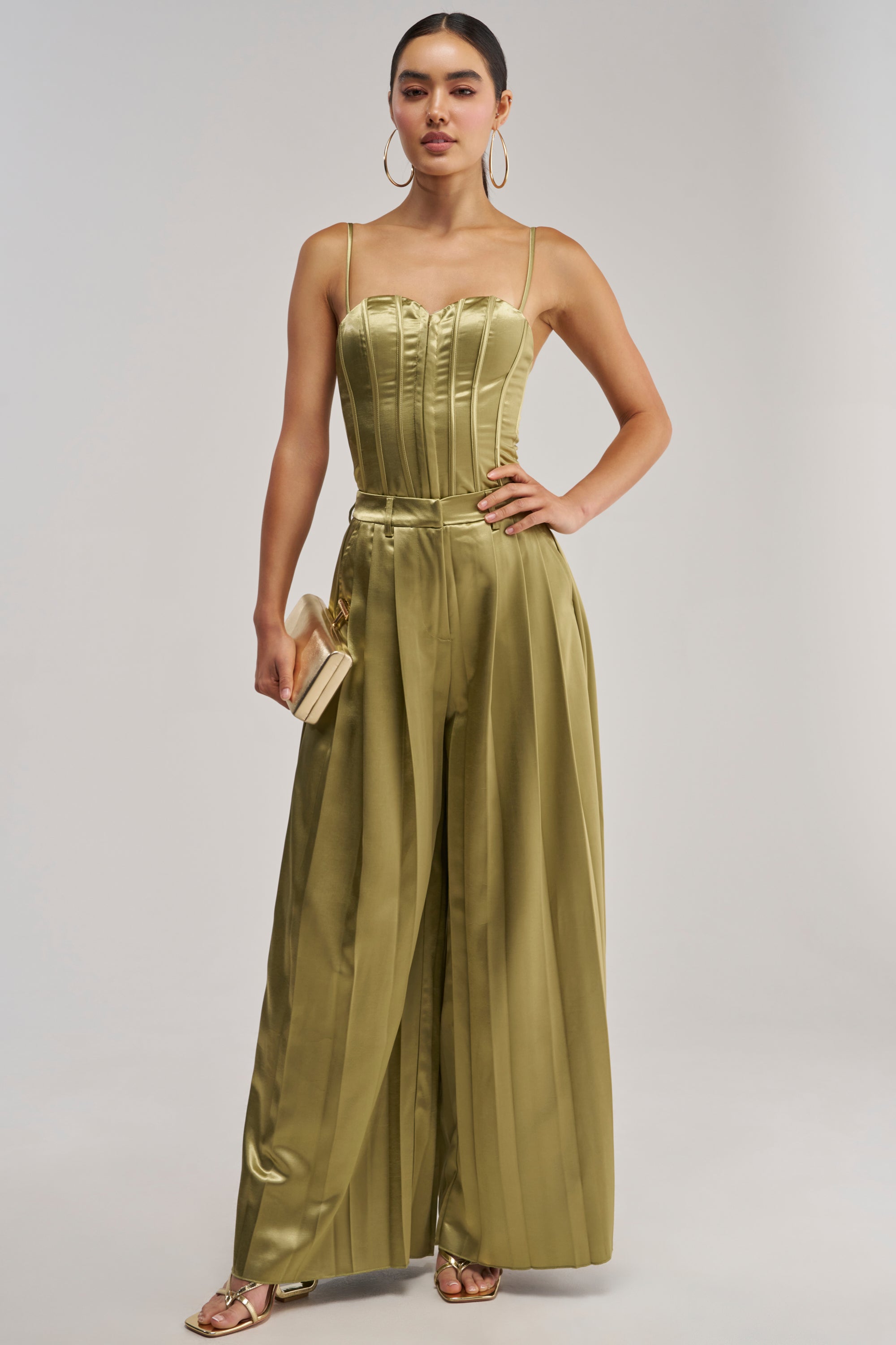 A woman stands against a plain background, wearing the IRIS SATIN PLEATED PANT IN OLIVE—a shiny olive green satin wide-leg trouser—paired with a matching spaghetti strap top, accessorized with hoop earrings, a small clutch, and open-toe heels.