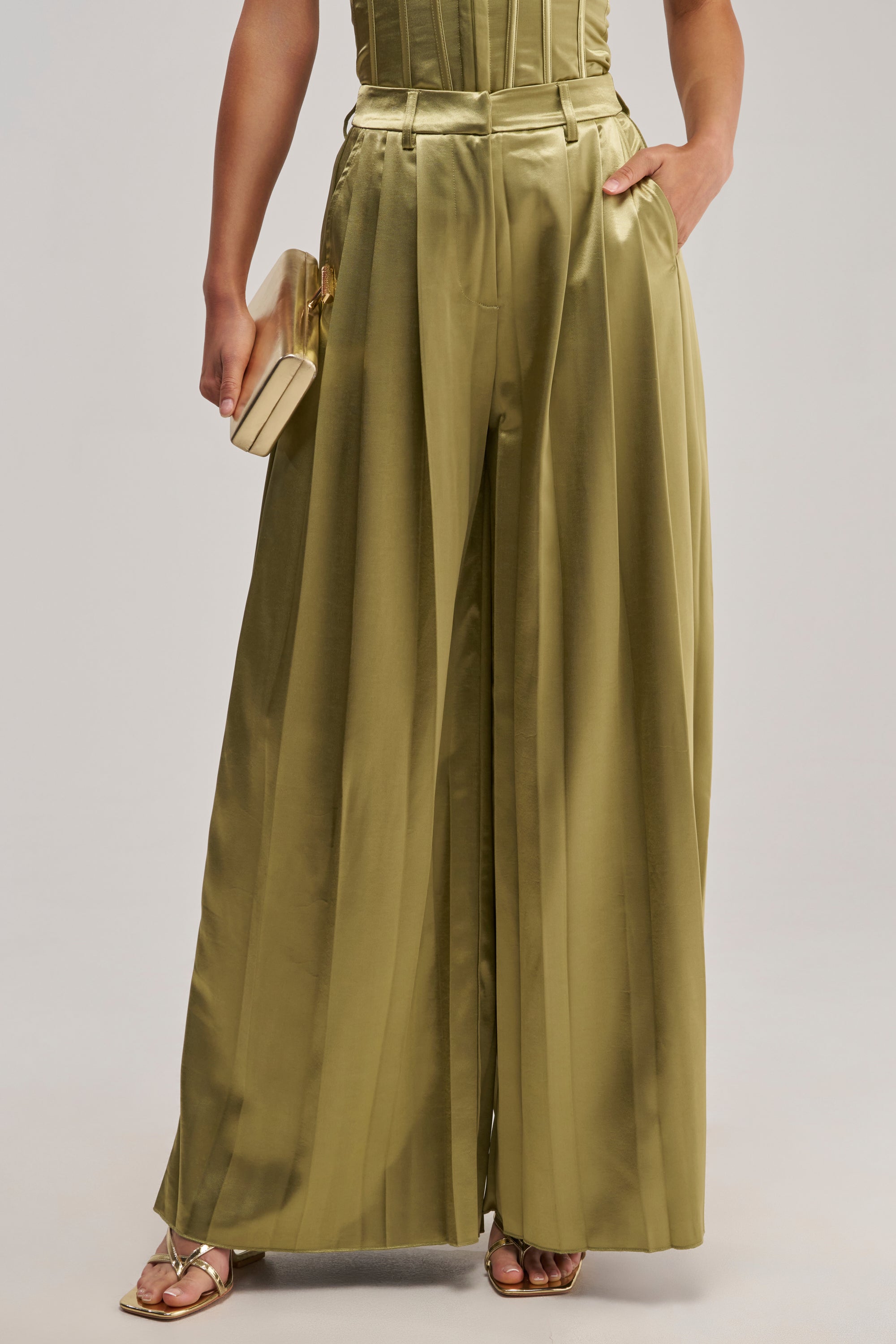 Wearing the IRIS SATIN PLEATED PANT IN OLIVE, this person styles the high-waisted, wide-leg trousers with a beige clutch in one hand and gold open-toe heels, with their upper body partially visible and one hand in a pocket.