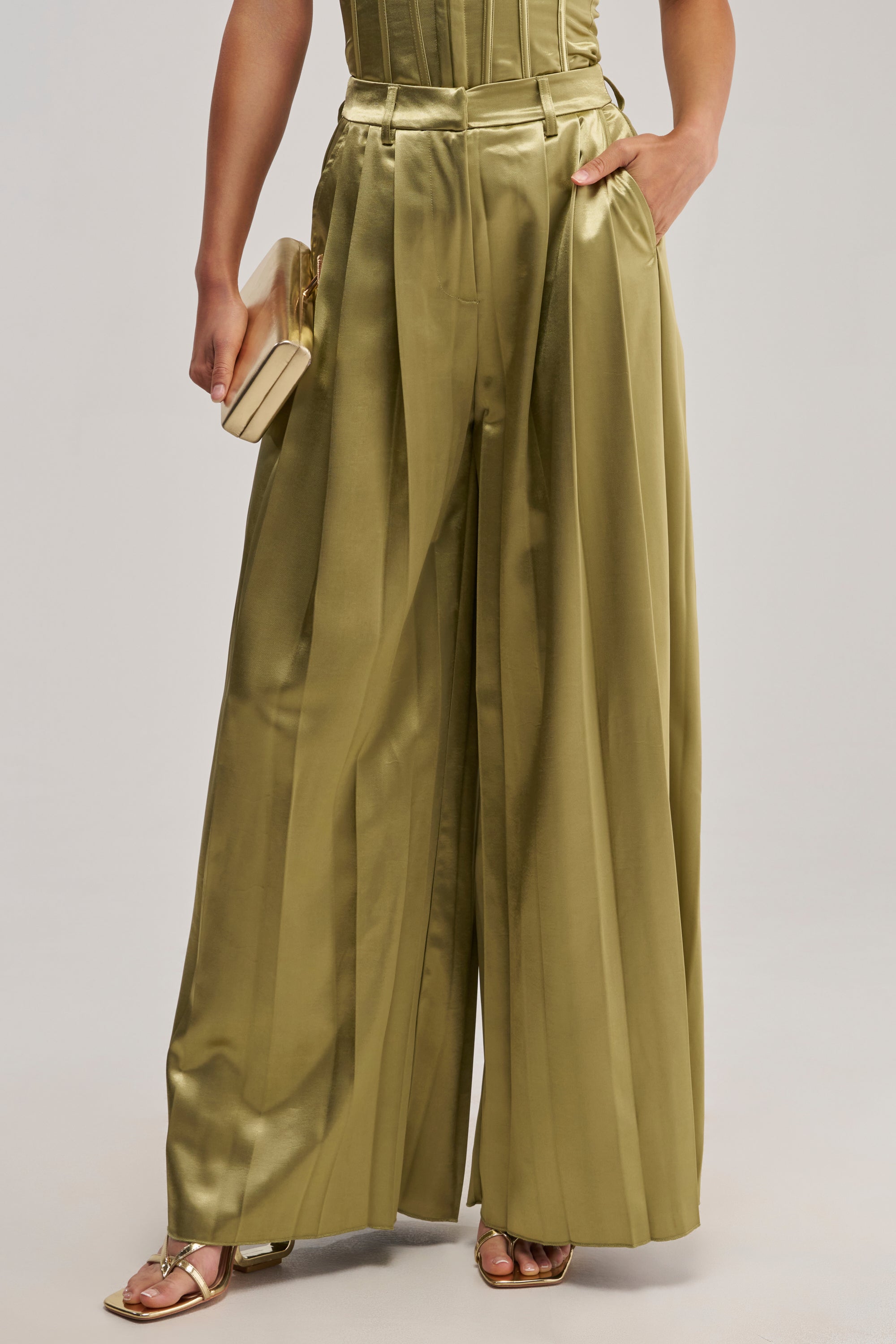 Wearing the IRIS Satin Pleated Pant in Olive, styled with a small beige clutch and strappy gold sandals.