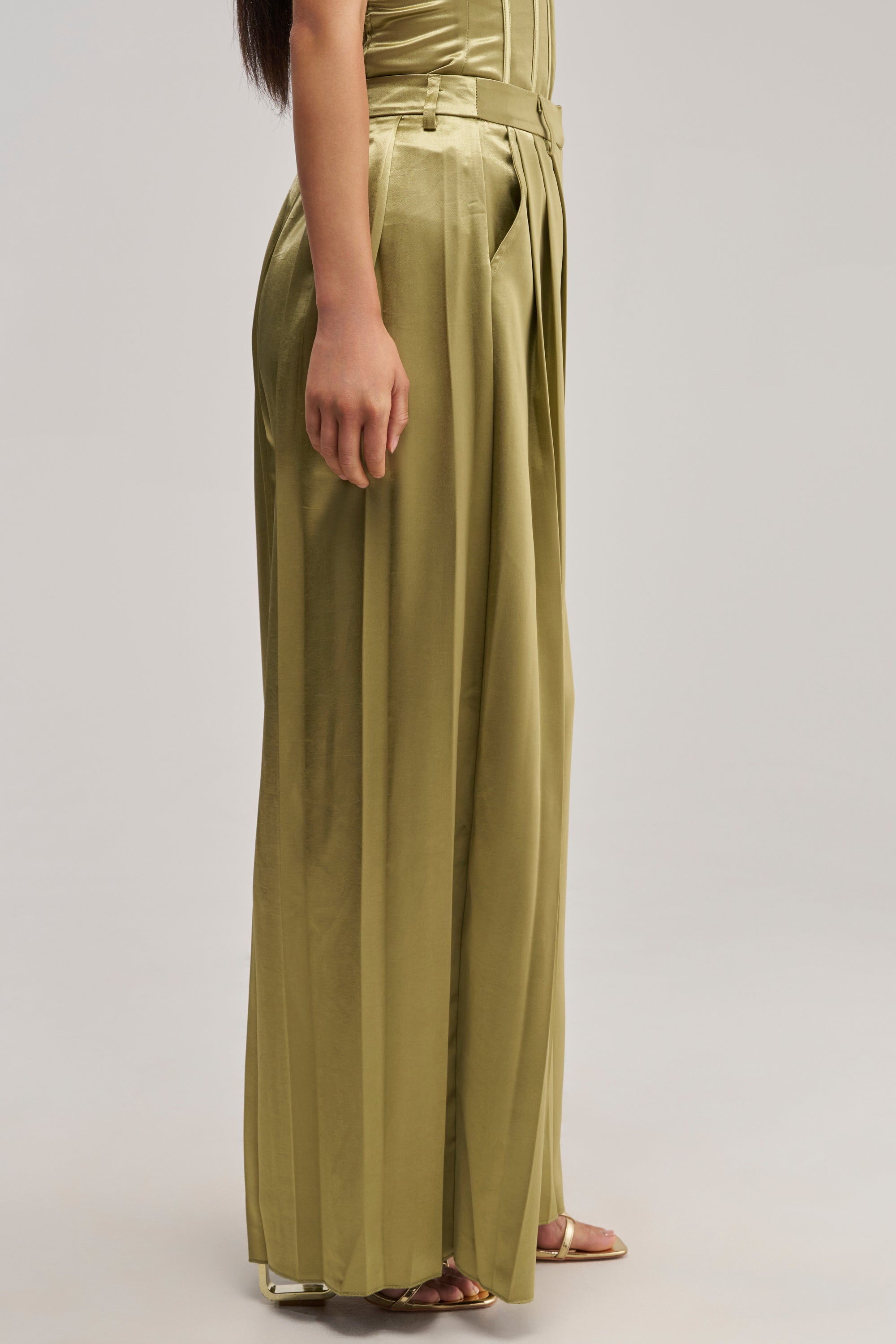 A person wearing the IRIS SATIN PLEATED PANT IN OLIVE styled with open-toe sandals stands against a plain light background, with only their lower body and one arm visible.