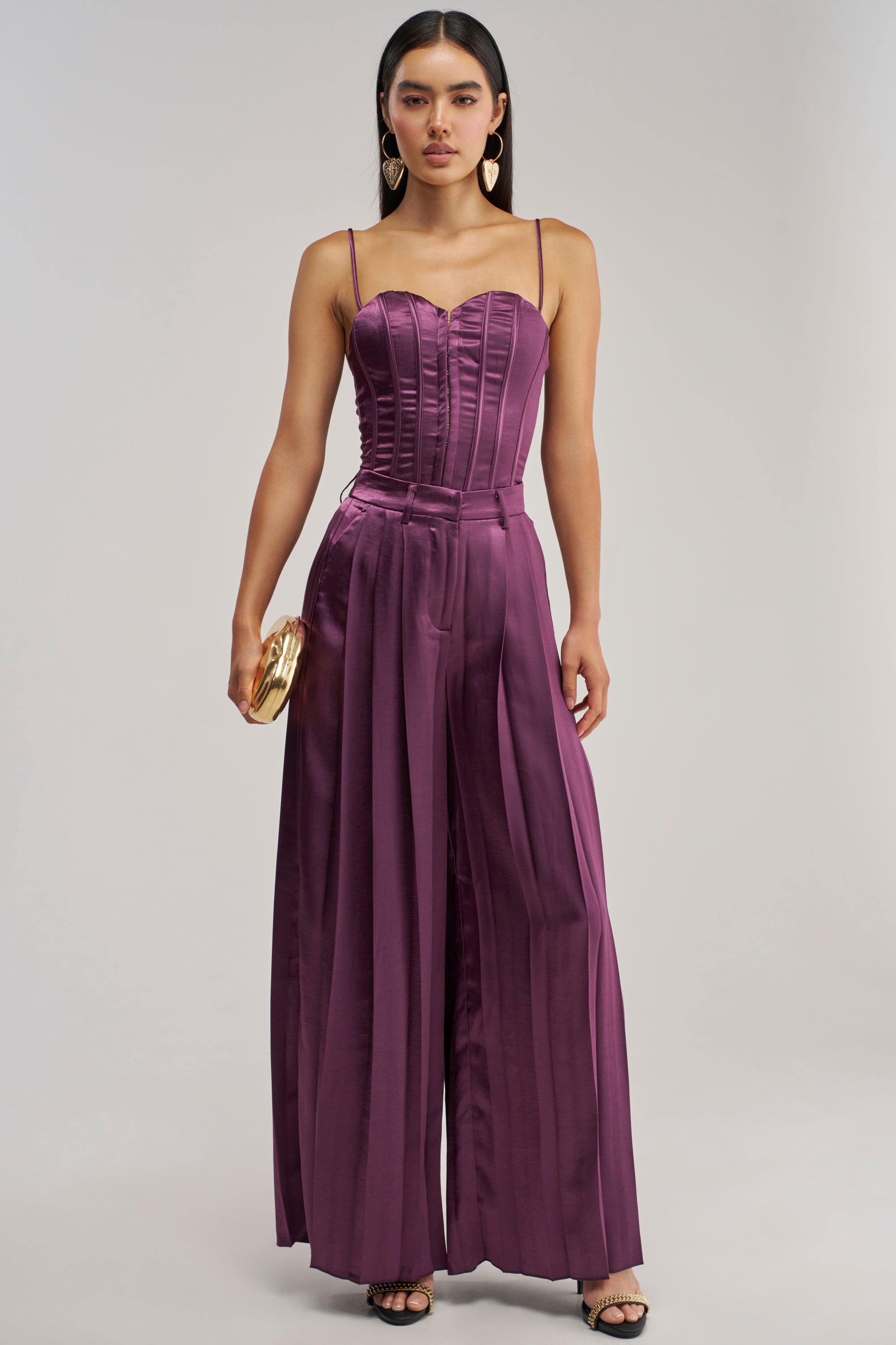 Wearing the IN TOO DEEP SATIN TROUSER, a woman dons a sleeveless, purple satin jumpsuit featuring wide-leg trousers and a structured bodice. Ideal for evening wear, she finishes the ensemble with a small gold clutch, hoop earrings, and black heels.