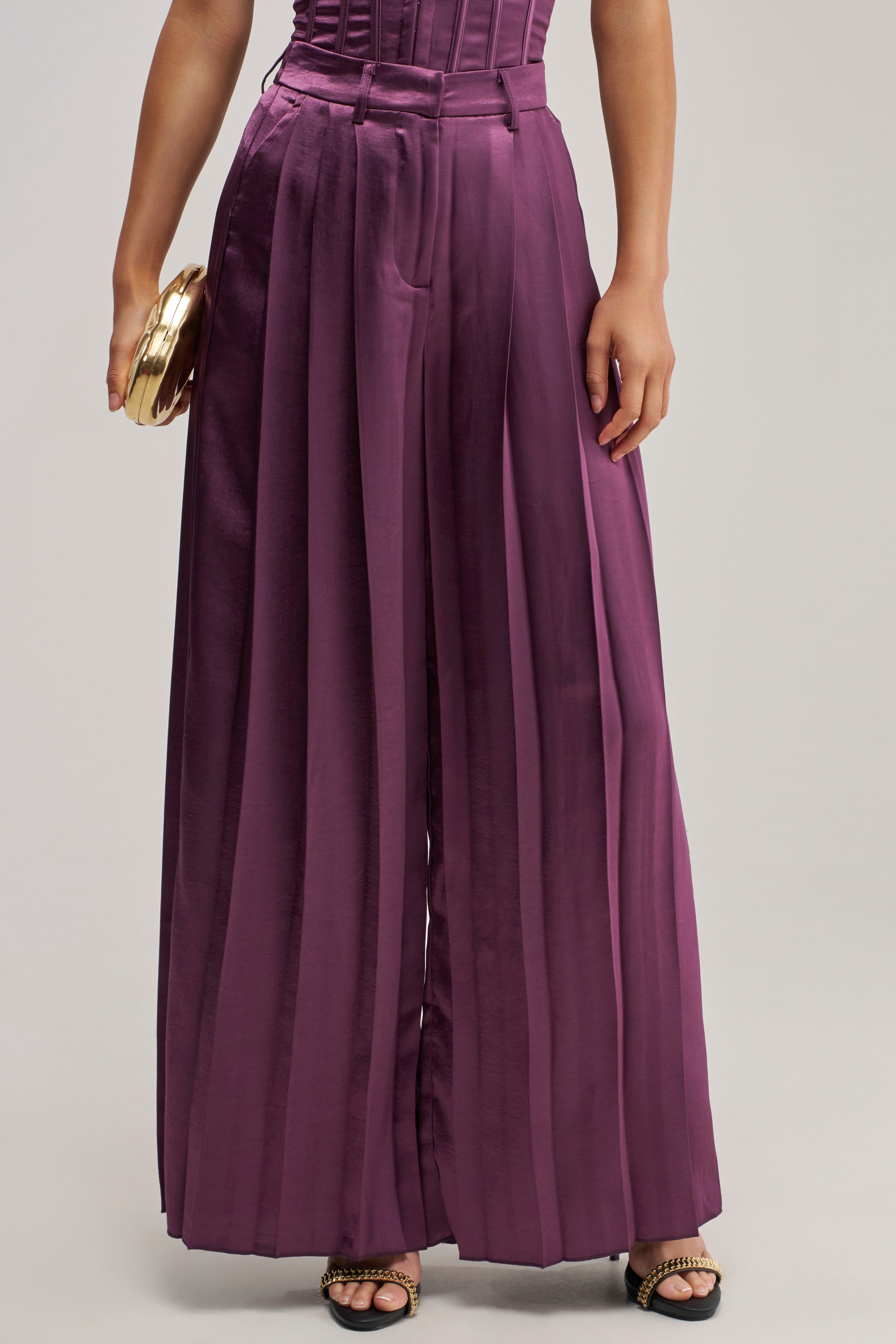 Wearing the IN TOO DEEP SATIN TROUSER—high-waisted, wide-leg plum satin pants—paired with a small round gold clutch and embellished black open-toe sandals creates an effortlessly chic look perfect for evening wear.