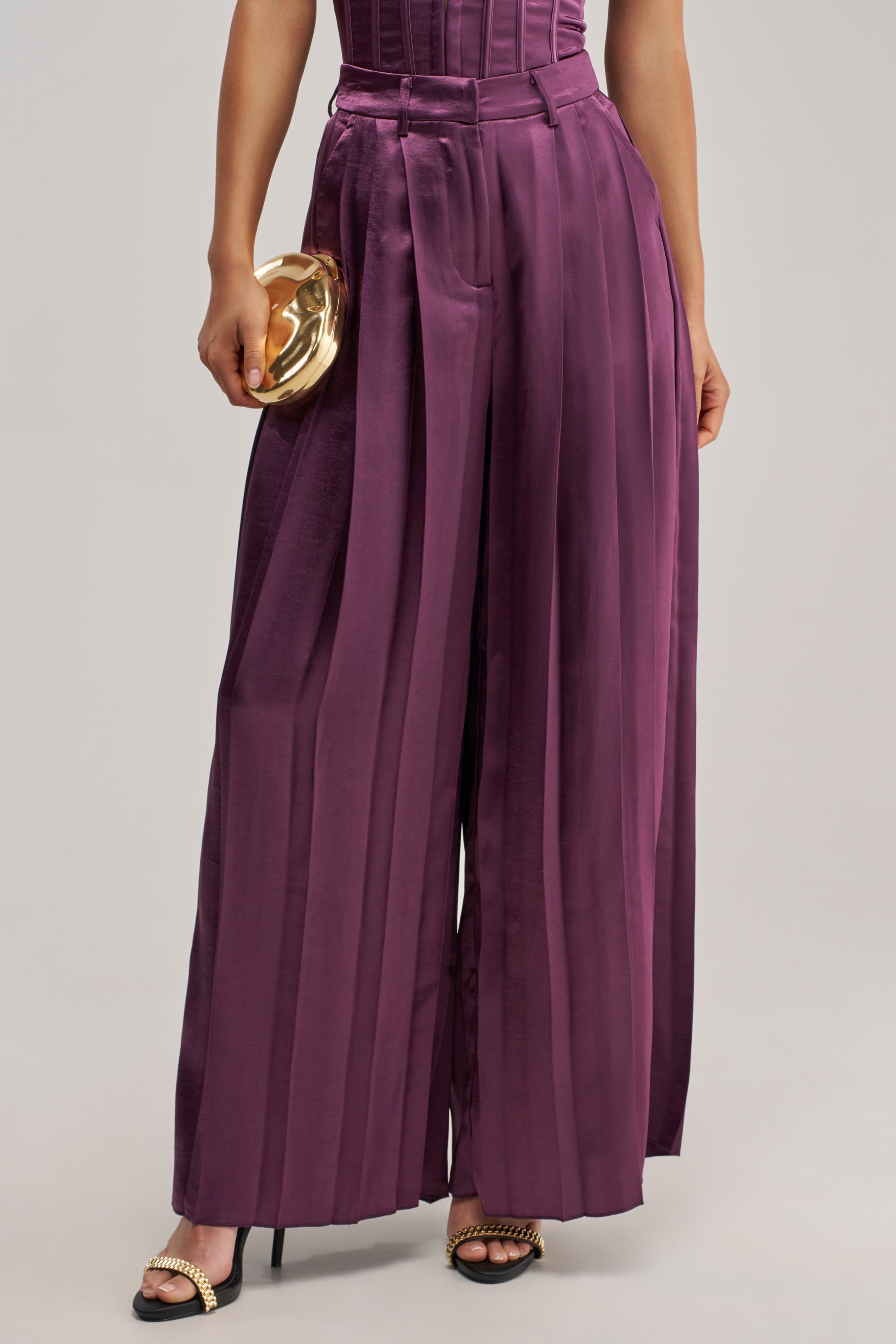 Wearing the IN TOO DEEP SATIN TROUSER—a high-waisted, wide-leg style in plum satin that’s perfect for evening wear—the person holds a small gold clutch and pairs the look with black heels featuring gold accents.