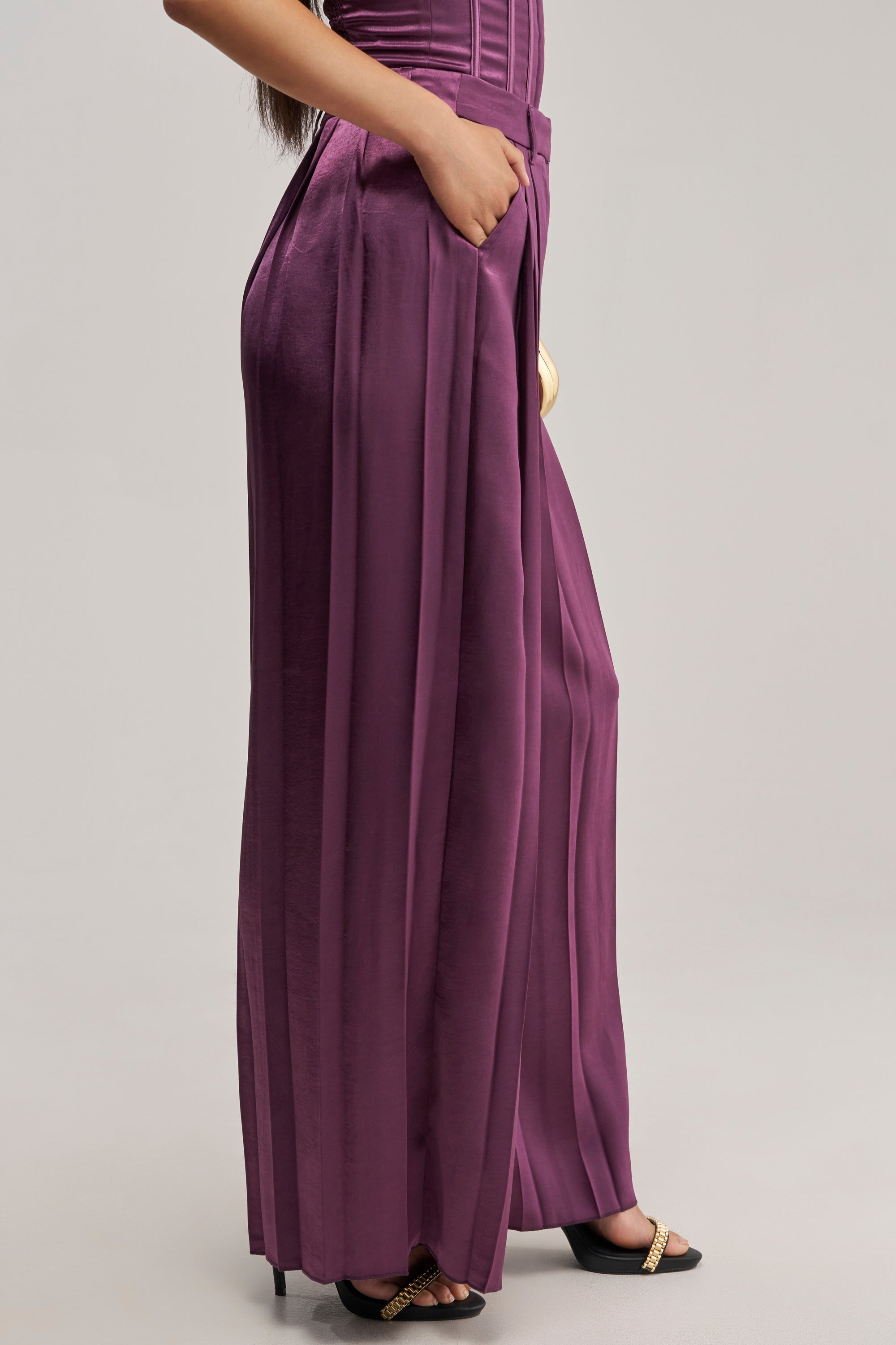 Wearing the IN TOO DEEP SATIN TROUSER in a high-waisted, wide-leg plum satin style paired with black heeled sandals featuring gold accents, this look against a plain light background exudes effortless chic—an ideal choice for sophisticated evening wear.