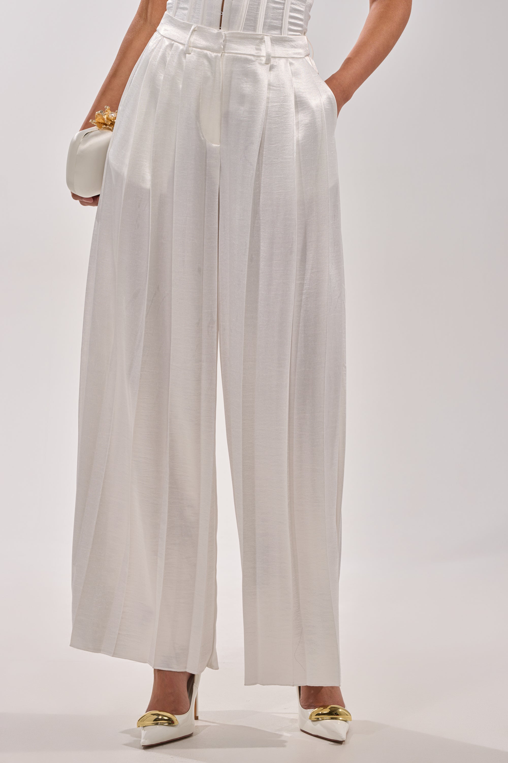 A person wearing the IRIS SATIN PLEATED PANT—high-waisted, wide-leg white satin trousers—paired with white heels featuring gold accents and a small white clutch; only the lower body is shown against a plain background. 0