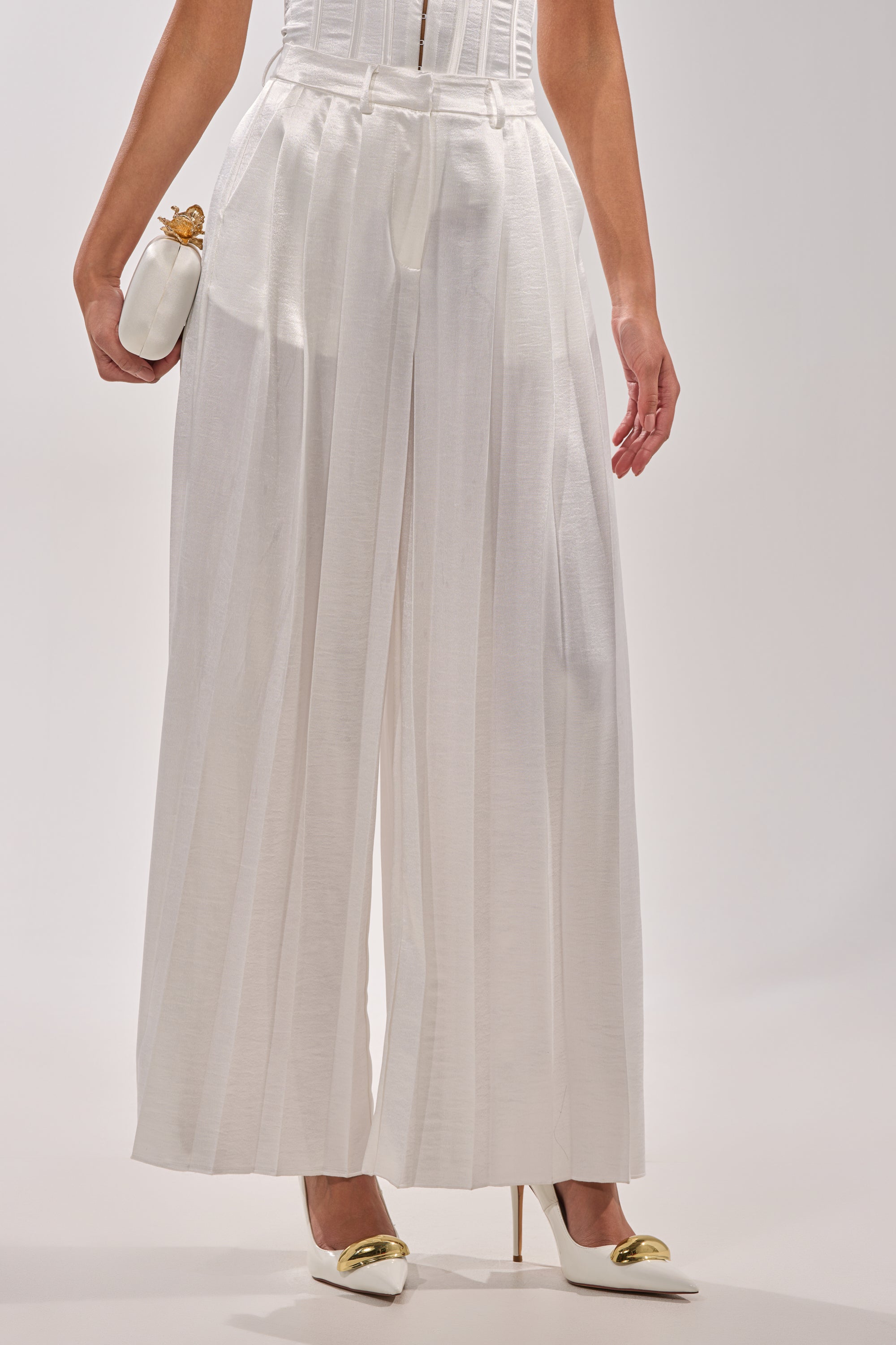 A person wears the IRIS SATIN PLEATED PANT—high-waisted, wide-leg white satin trousers—with white pointed-toe heels featuring gold accents and holds a small white clutch against a plain background. 1