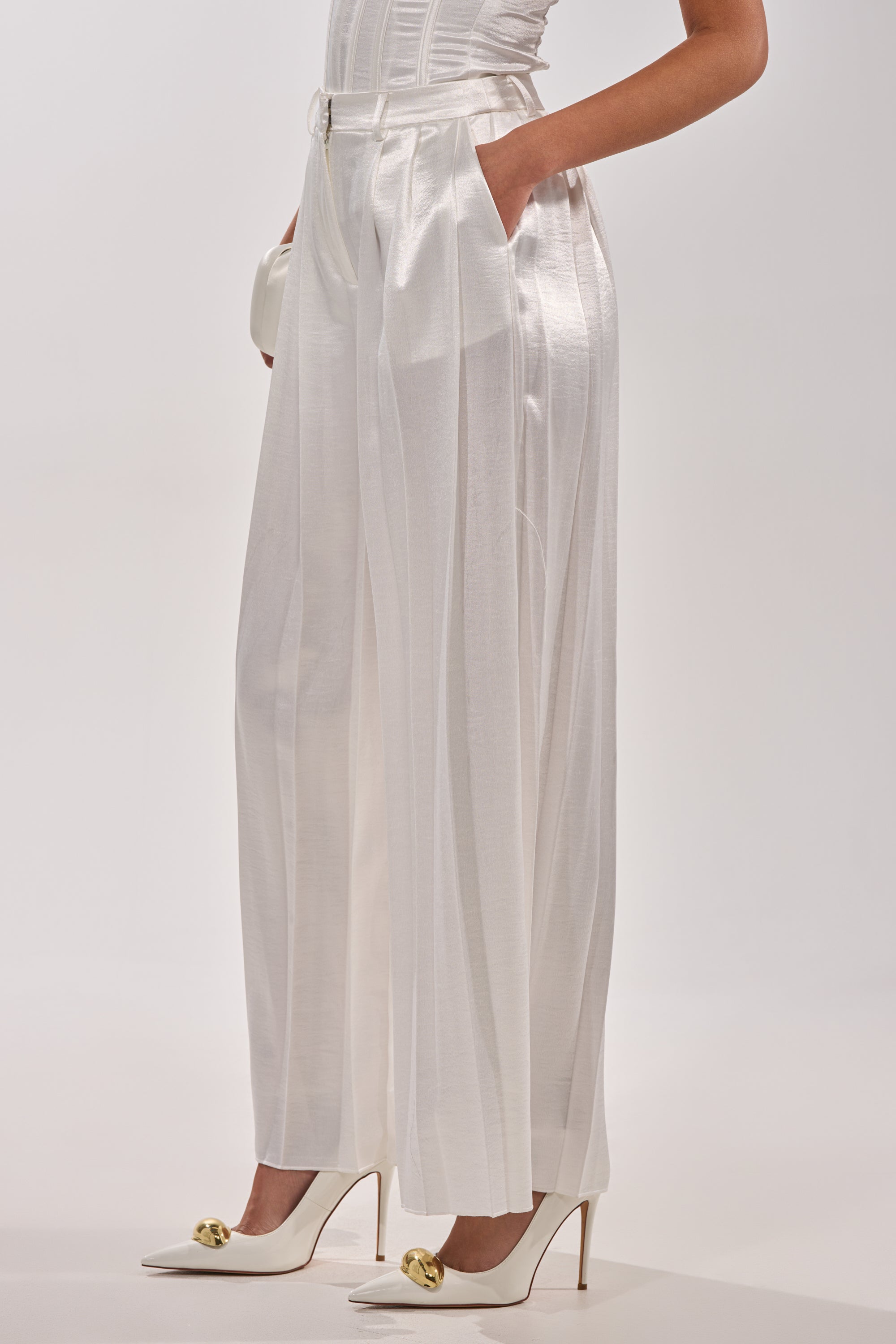 Wearing the IRIS SATIN PLEATED PANT, a high-waisted, wide-leg white satin trouser with pleats, paired with white heeled shoes with gold details and a fitted top. Their hands rest casually in the pant’s pockets. 2