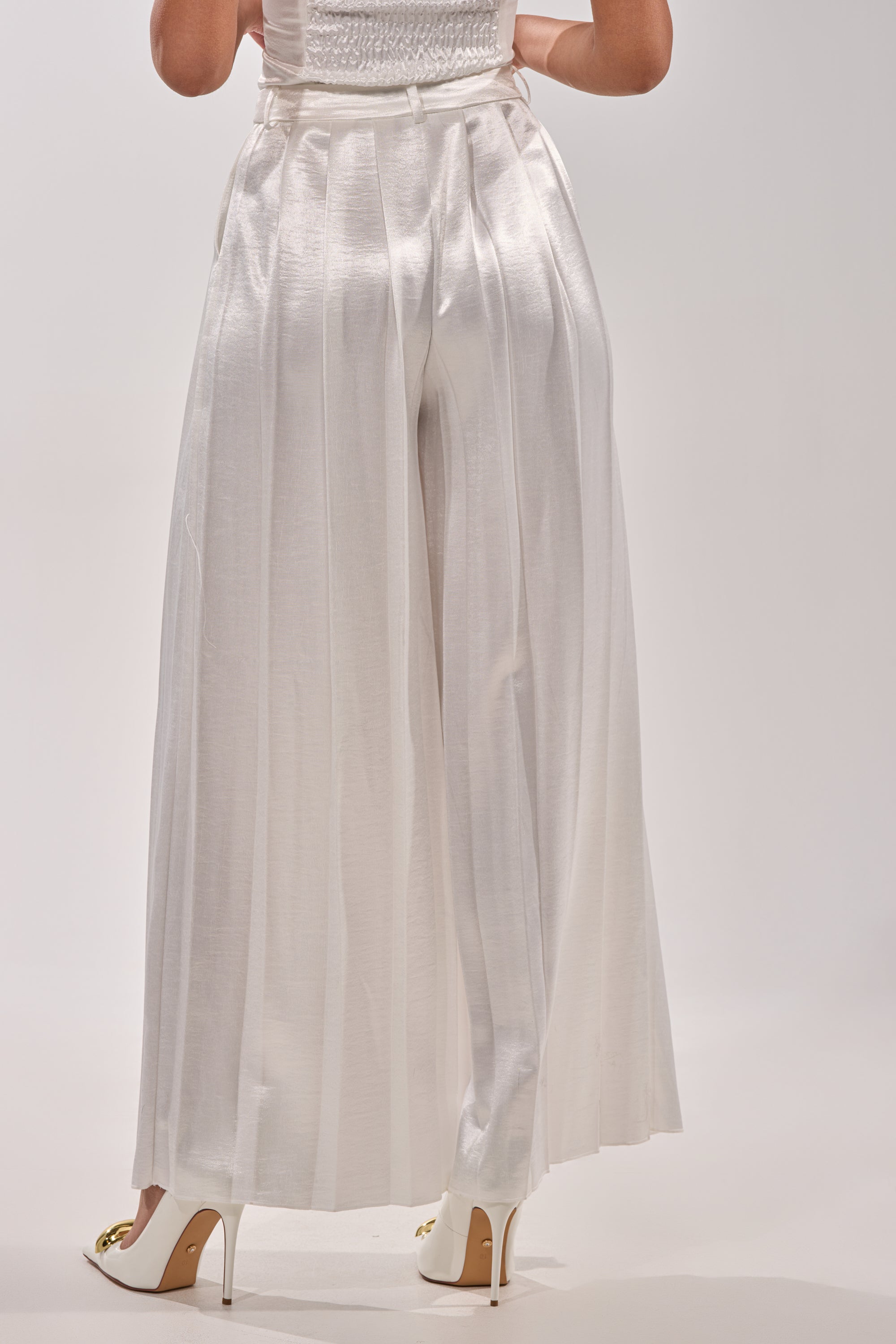 Showcasing minimalist chic, a person is seen from the back in IRIS SATIN PLEATED PANT paired with white high-heeled shoes against a plain light background. 3