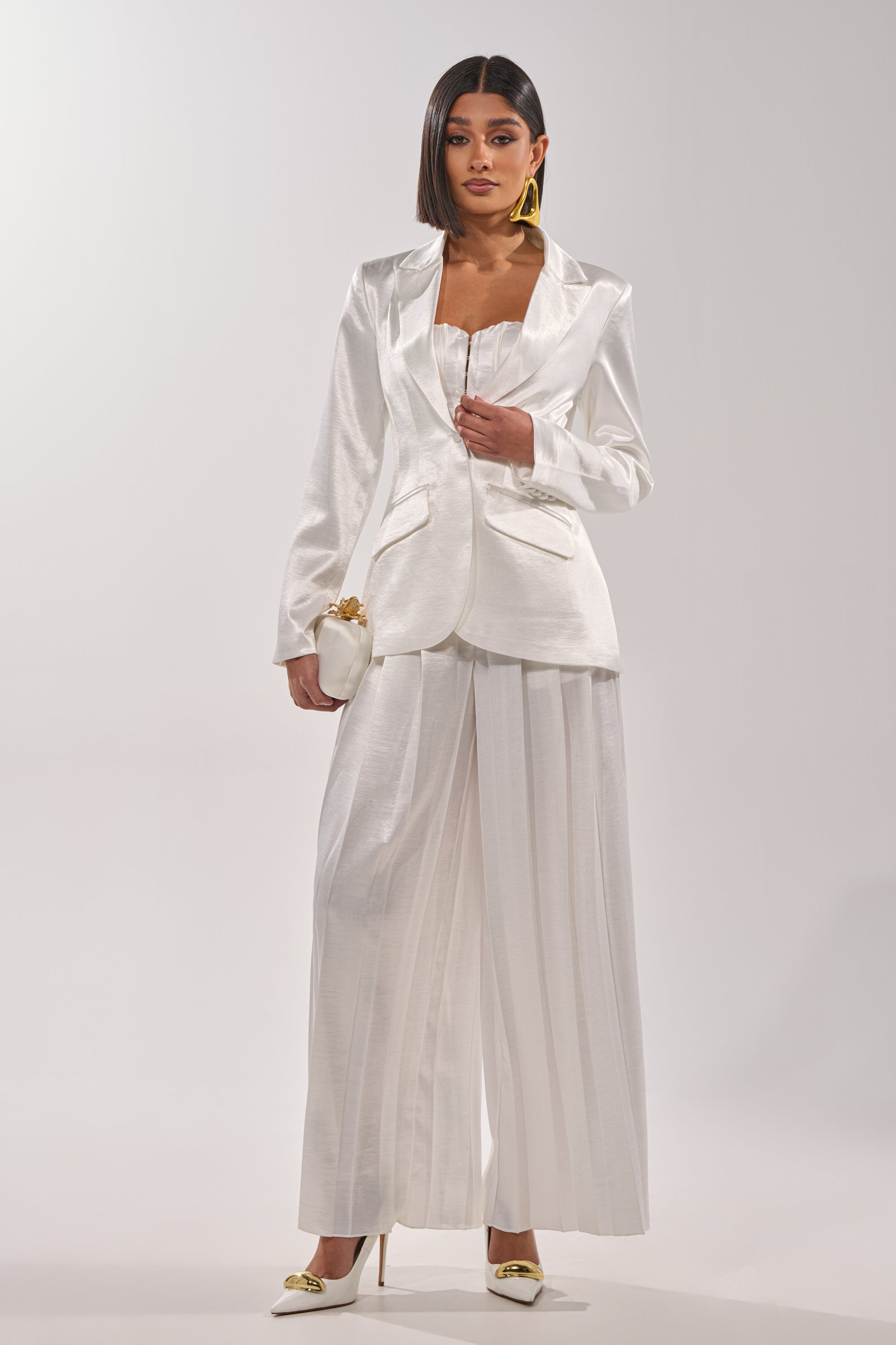 A woman poses in the IRIS SATIN PLEATED PANT, a chic high-waisted wide-leg white design, styled with a fitted blazer, matching top, gold pointed heels, statement yellow earrings, a small clutch, and straight shoulder-length hair. 5