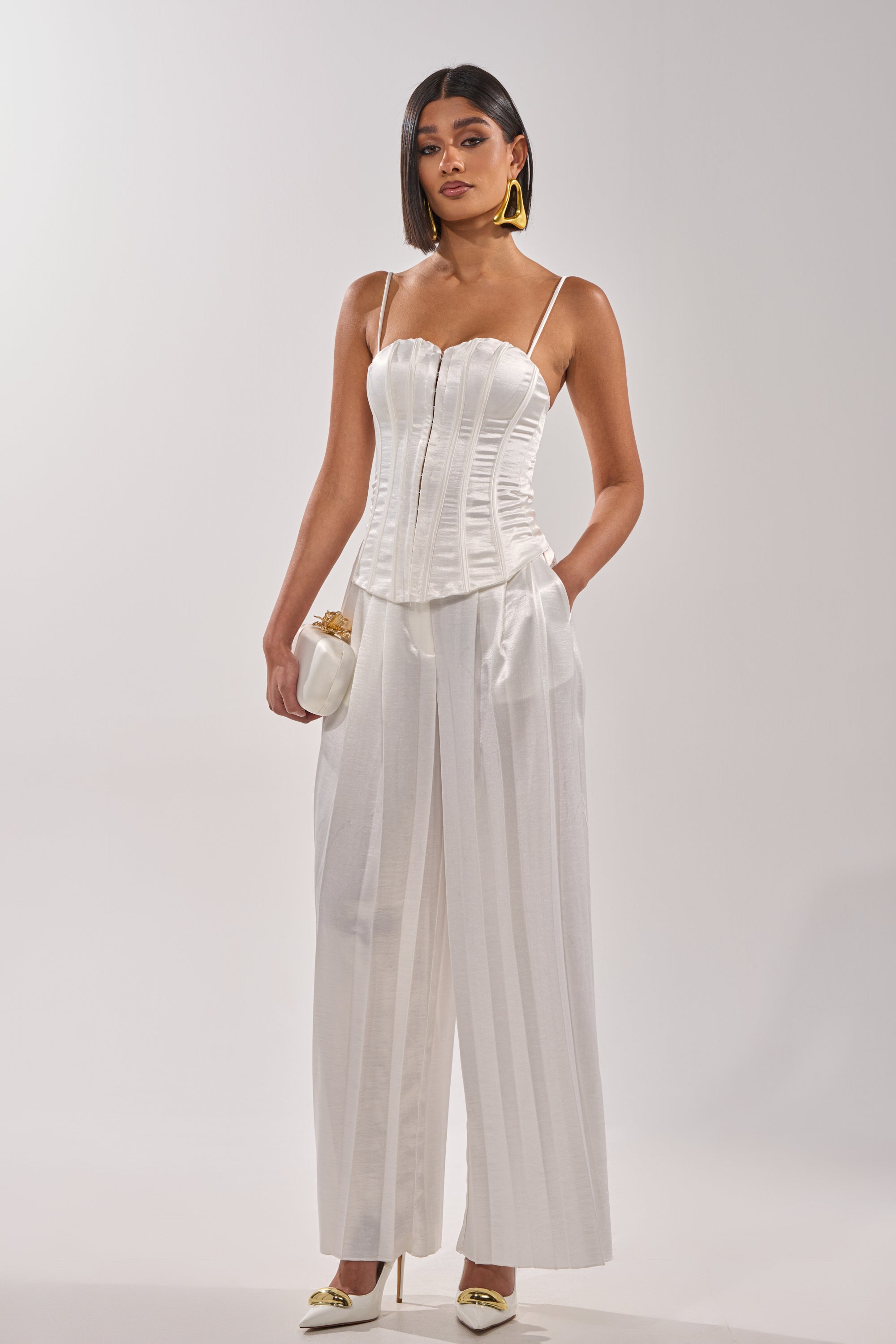 A woman with straight dark hair and gold earrings stands against a plain background wearing the IRIS SATIN PLEATED PANT, a white satin corset top, cream heels, and holds a small white clutch for a luxe minimalist chic look. 6