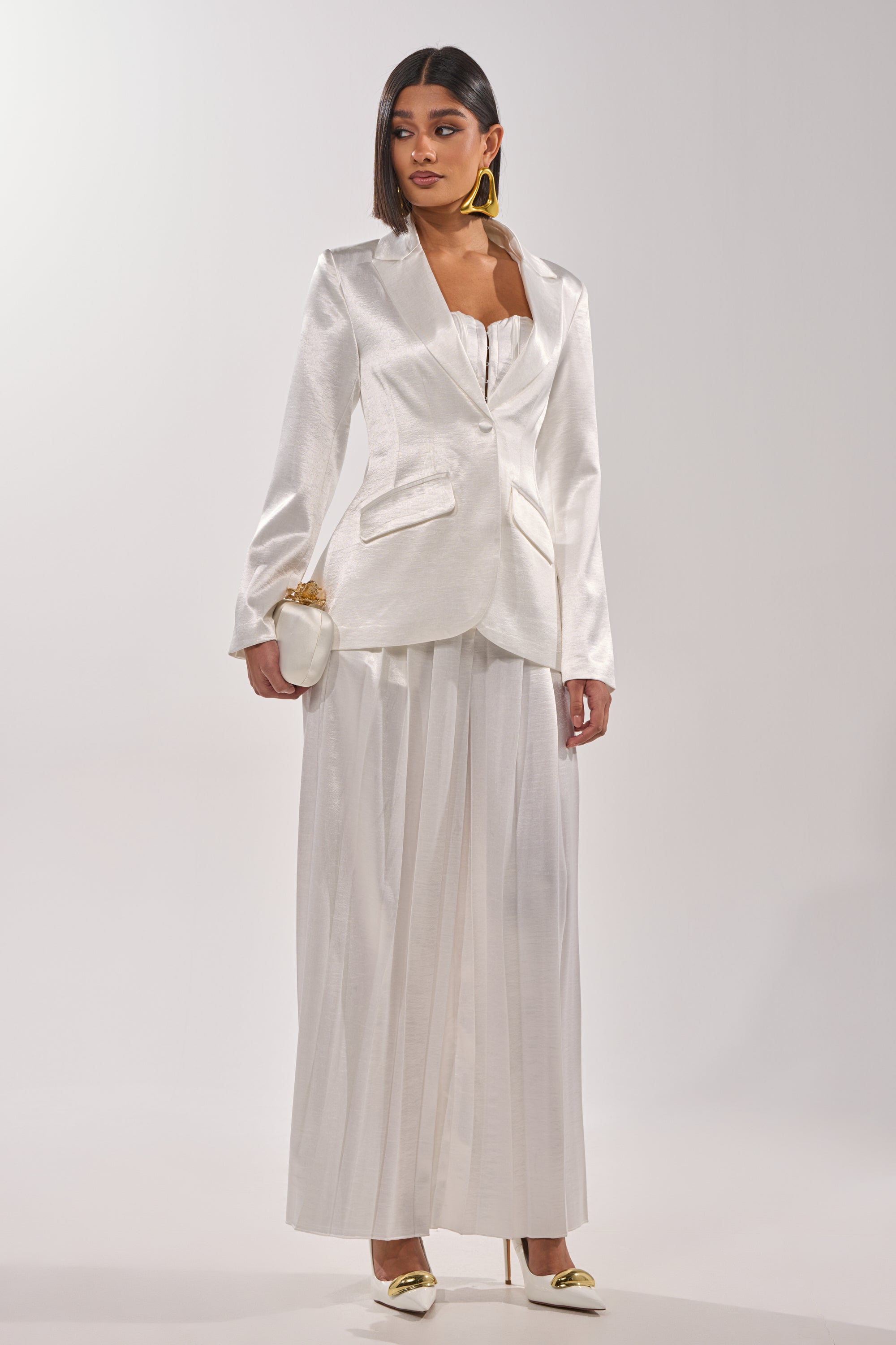 A woman poses against a plain background wearing IRIS SATIN PLEATED PANT, a shiny white blazer, gold heels, bold earrings, and holds a small white clutch while gazing slightly to the side. 7
