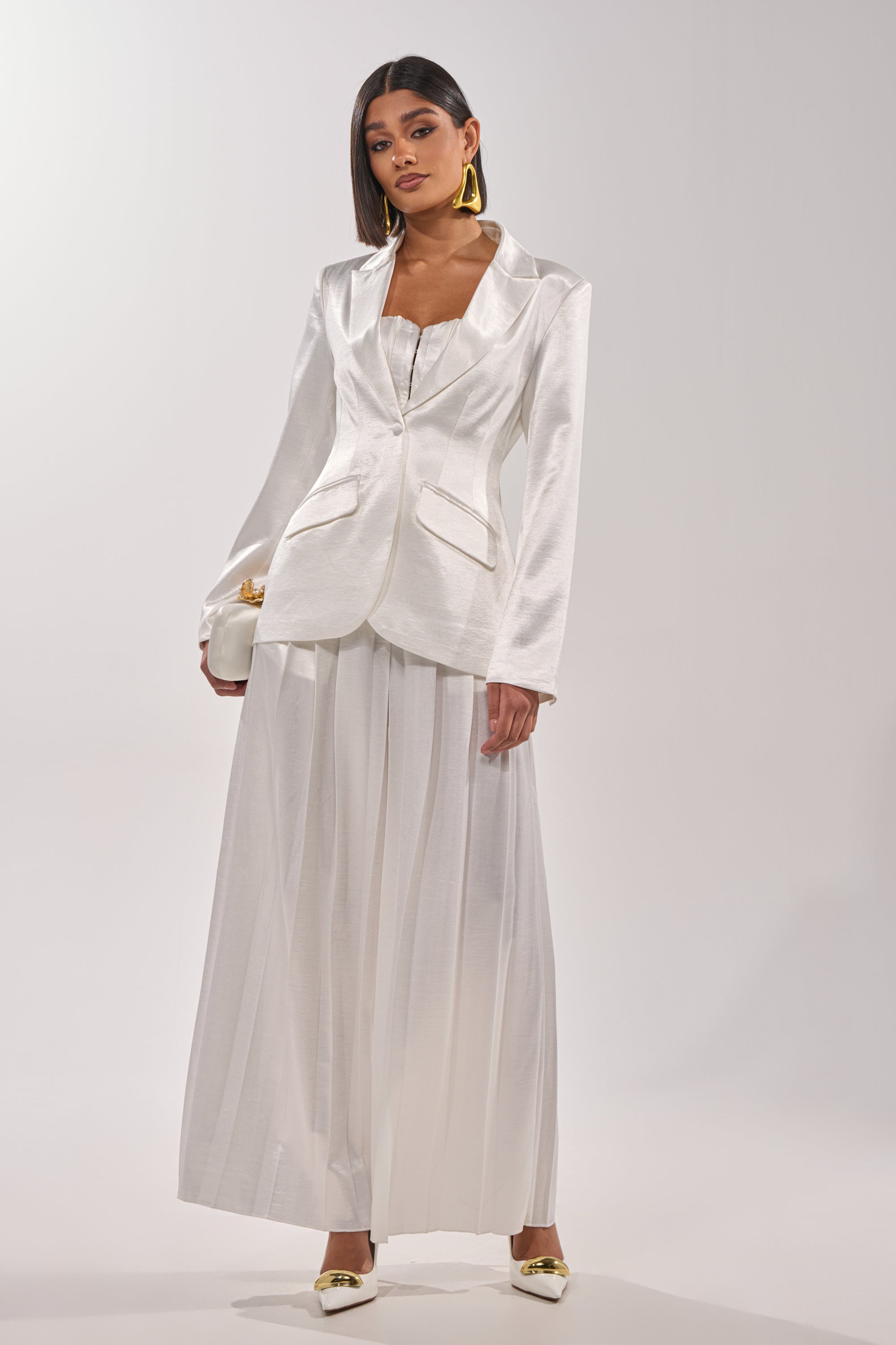 A woman poses against a plain backdrop wearing the IRIS SATIN PLEATED PANT with a white satin blazer, pointed-toe heels, large gold earrings, and a sleek bob hairstyle. 8