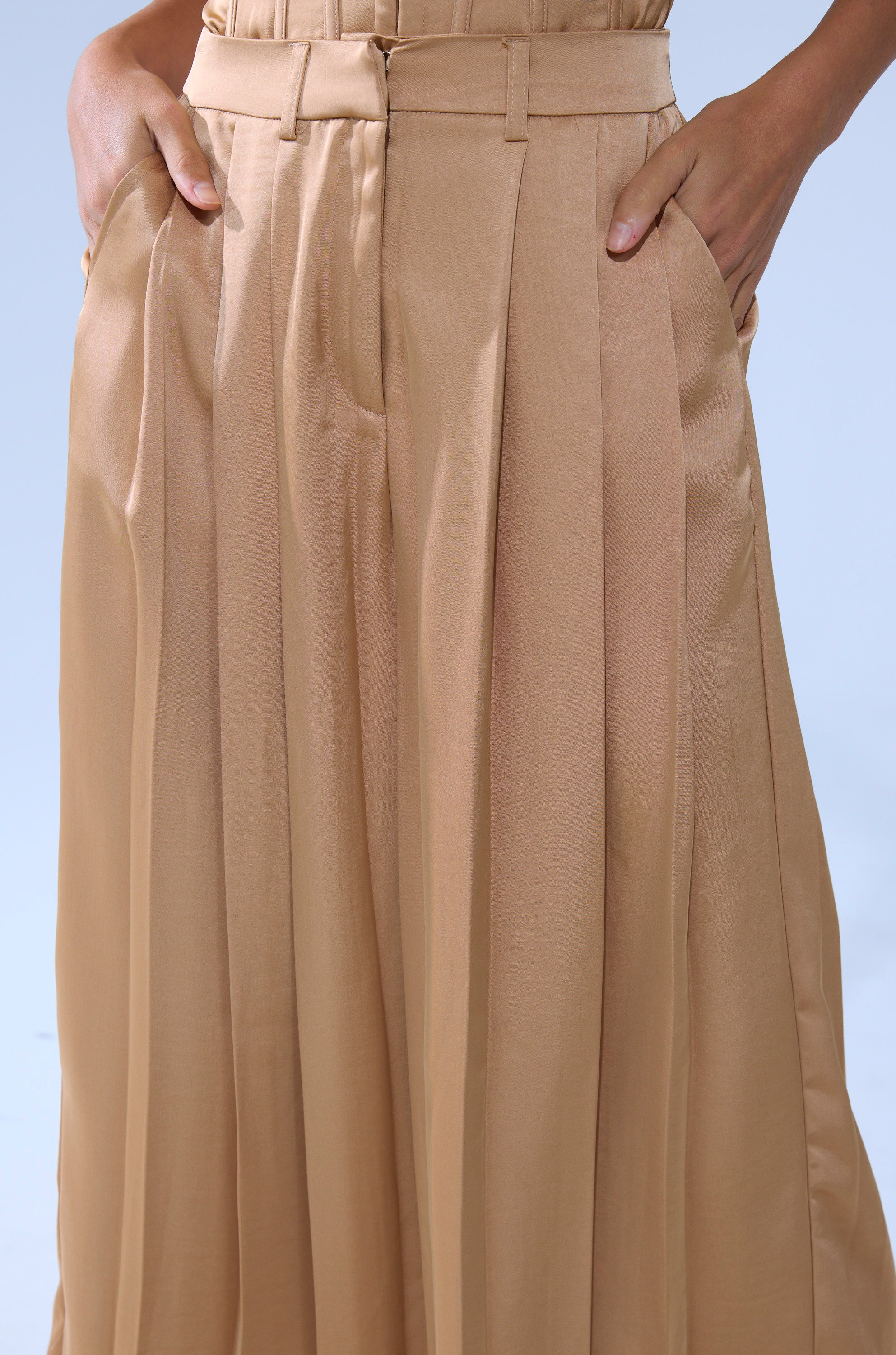 Someone wearing HANSEL MOCHA SATIN PALAZZO PANT, featuring a high-rise wide-leg design with pleats and hands in pockets, stands against a plain light blue background.