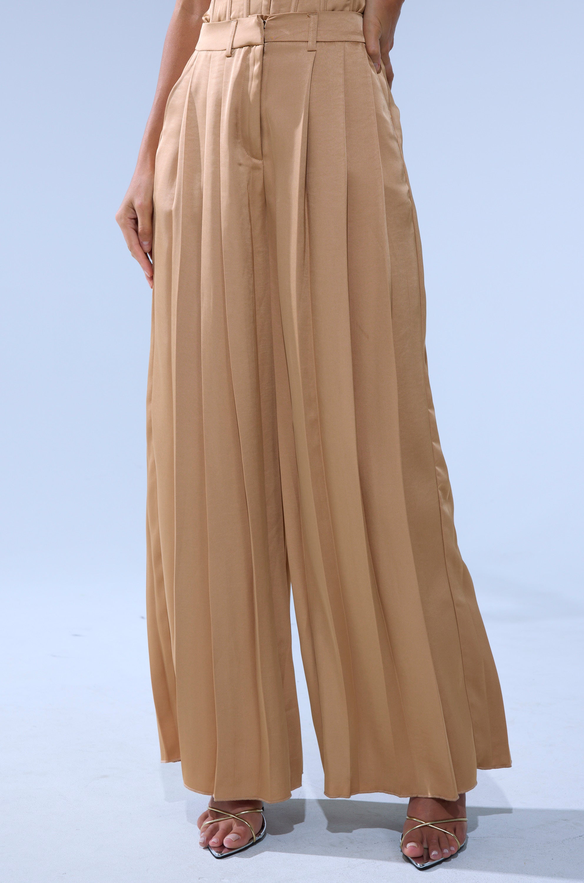A person wearing the HANSEL MOCHA SATIN PALAZZO PANT from AKIRA Label and silver open-toe heels stands against a light blue background.