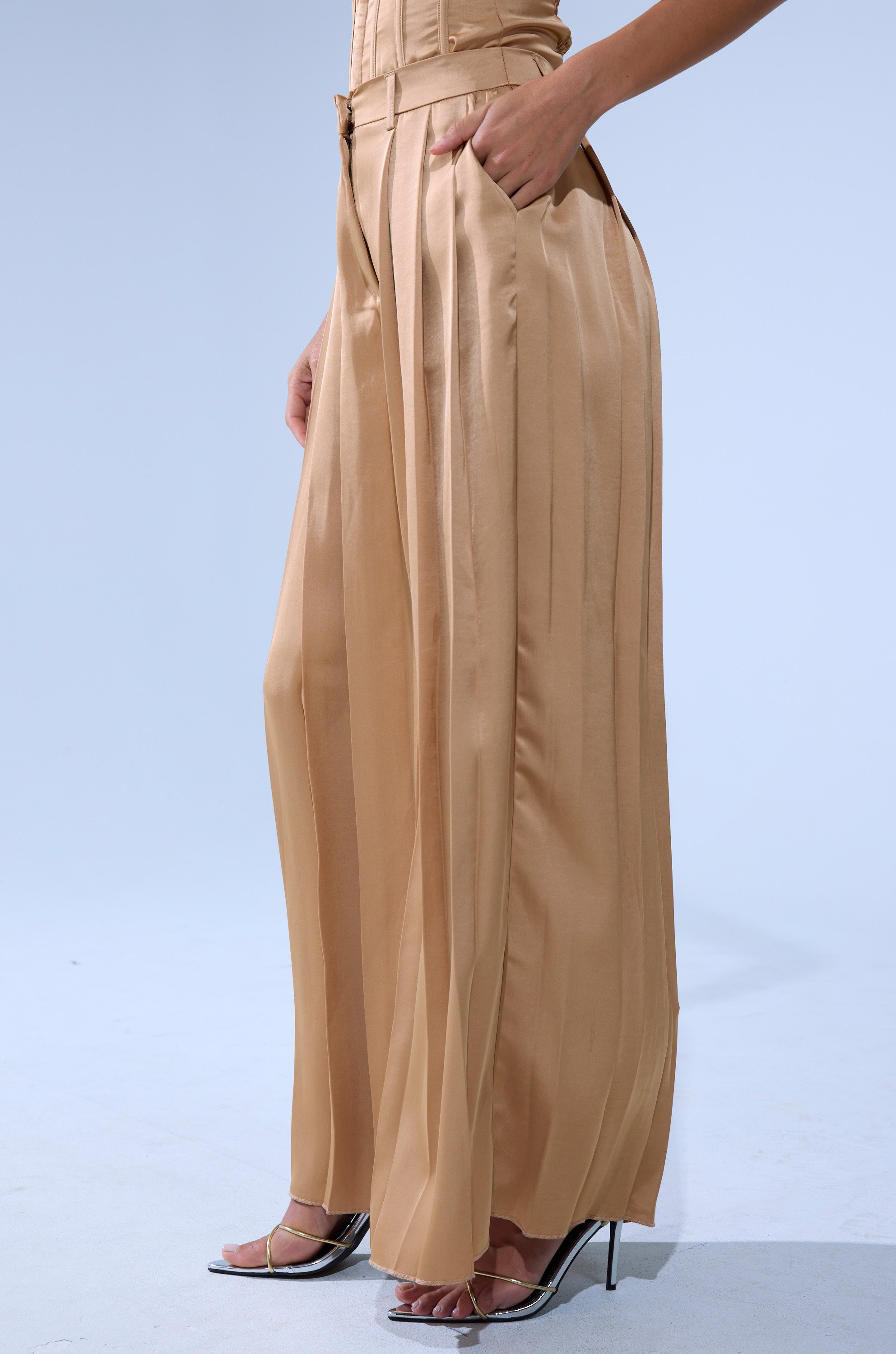 A person wears the HANSEL MOCHA SATIN PALAZZO PANT, featuring high-rise wide-leg pleated satin, paired with clear strap high heels. Standing against a plain light background, one hand rests in the pant pocket.