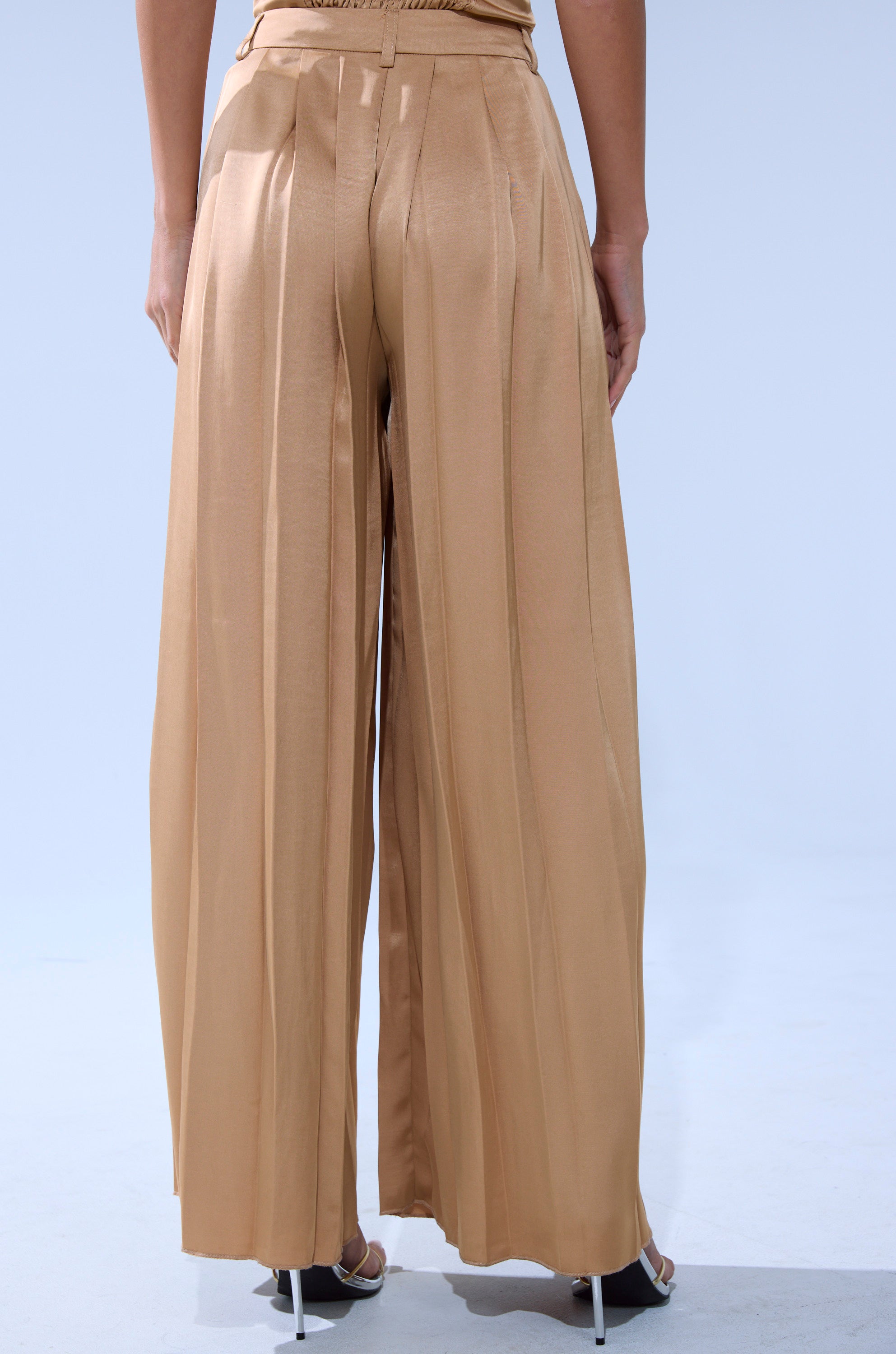 A person is wearing the HANSEL MOCHA SATIN PALAZZO PANT, shown from the back to showcase its high-rise, wide-leg design, vertical pleats, loose fit, and flowy satin fabric.