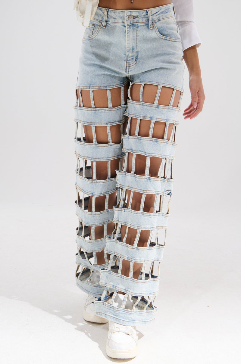 Wearing the CAGED WIDE LEG DENIM featuring large rectangular leg cutouts, this person styles it with white sneakers and a cropped top, standing against a plain white background.