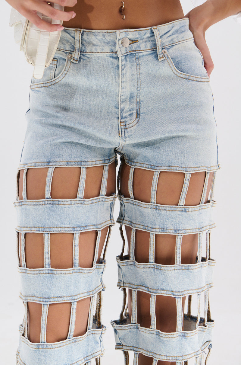 A person wears CAGED WIDE LEG DENIM in light blue, featuring large square cutouts along the legs that reveal skin, with a classic waistband and pockets. The person also has a belly button piercing.
