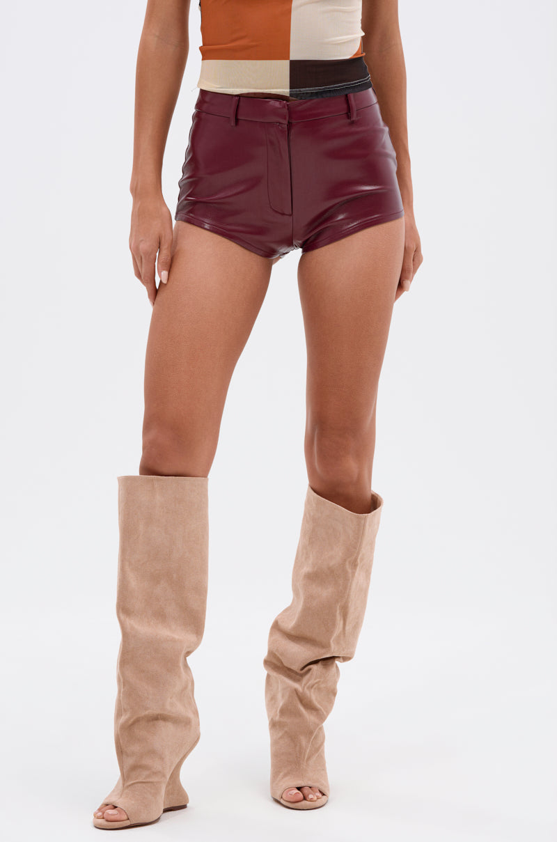 Wearing the OH HOT DAMN FAUX LEATHER HOT SHORT in burgundy, a color-blocked sleeveless top, and beige slouchy knee-high peep-toe boots, a person stands against a plain white background. 2