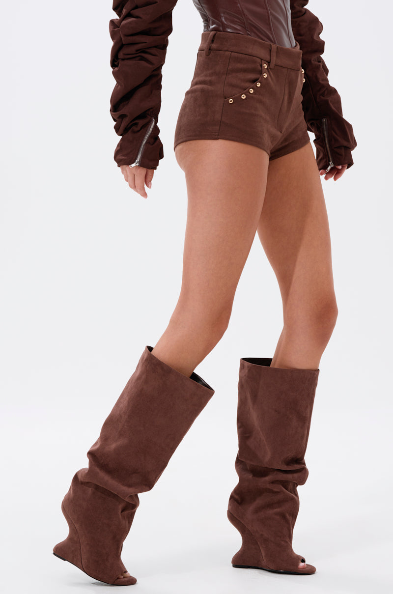 A person wears the TAYLOR SUEDE HOT SHORT in brown with gold buttons, paired with a long-sleeve brown top and tall slouchy brown wedge boots, standing against a plain white background. 3