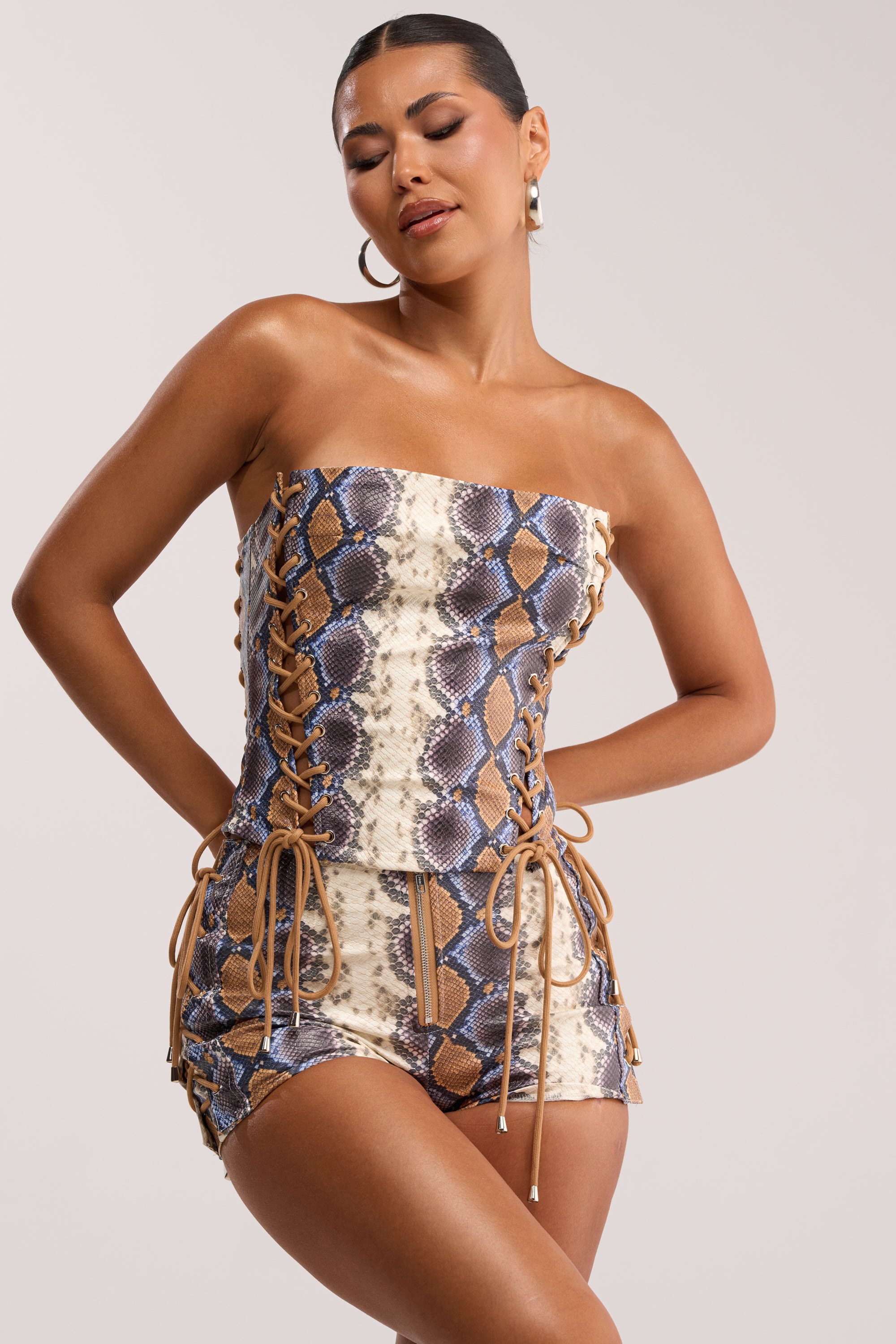 A woman models the YOU KNOW I WANT YOU BABY LACE UP CORSET, a strapless romper with a brown, beige, and blue snakeskin print and lace-up sides. She wears hoop earrings and has slicked-back hair, posing confidently against a plain background.
