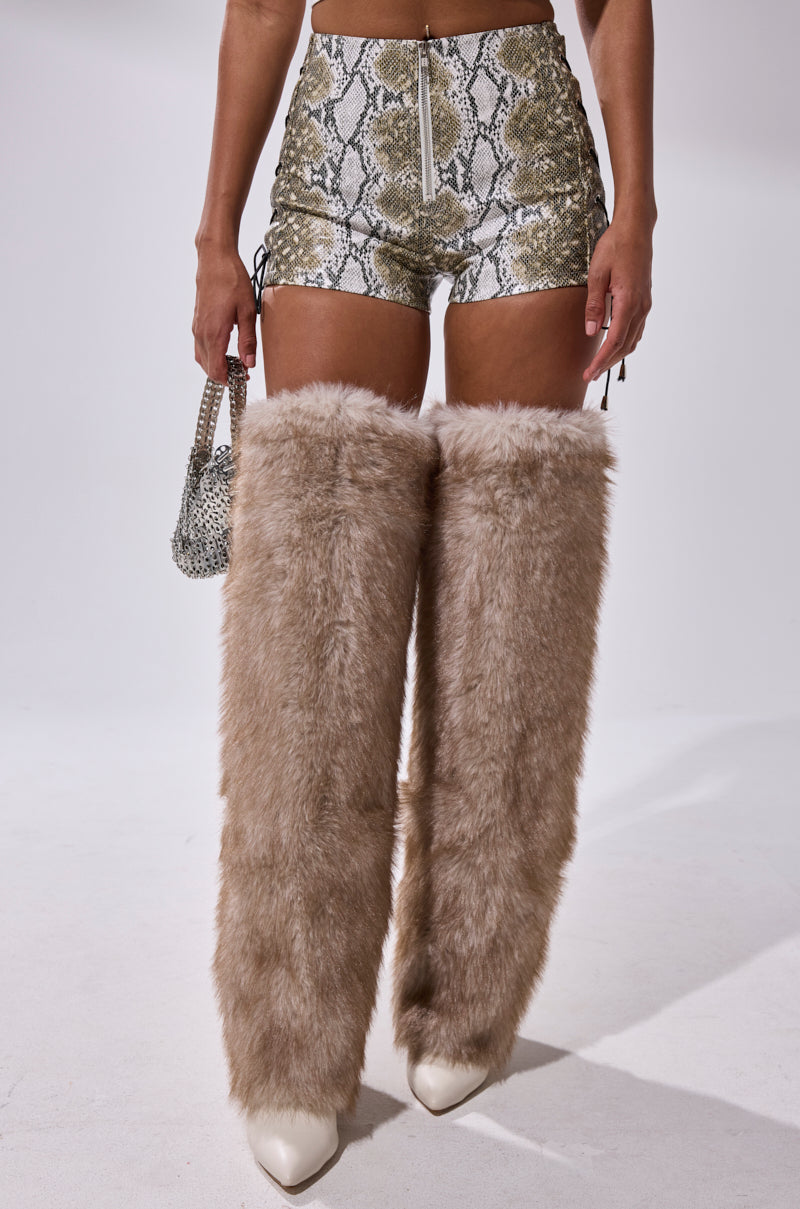 A person wearing the JUST HOT AND TALKING FAUX LEATHER SHORT WITH LACE UP, paired with beige faux fur leg warmers over white pointed boots and holding a silver handbag, stands against a plain white background. 2