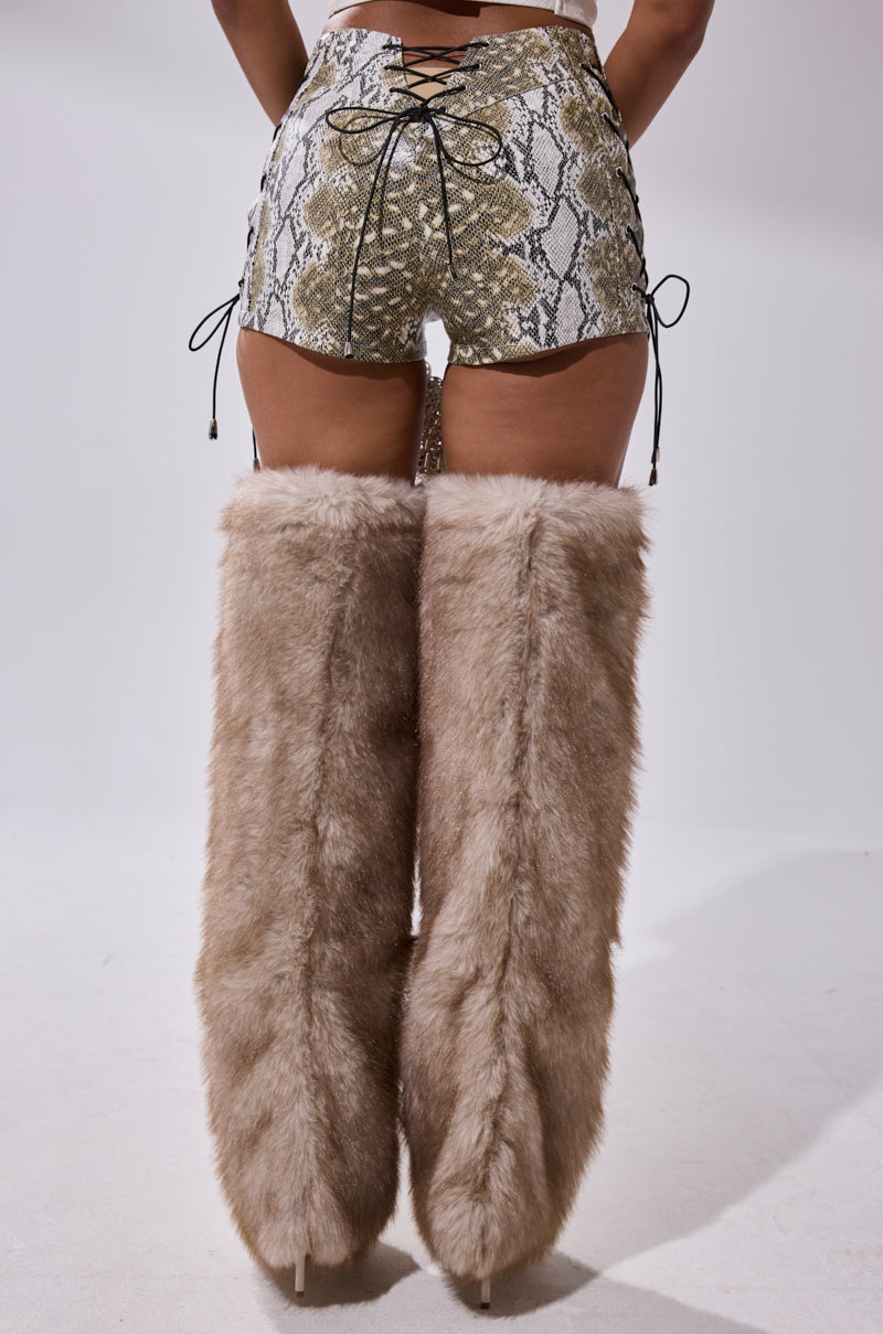 A person faces away, wearing the JUST HOT AND TALKING FAUX LEATHER SHORT WITH LACE UP and tall, tan faux fur thigh-high boots against a plain white background. 4