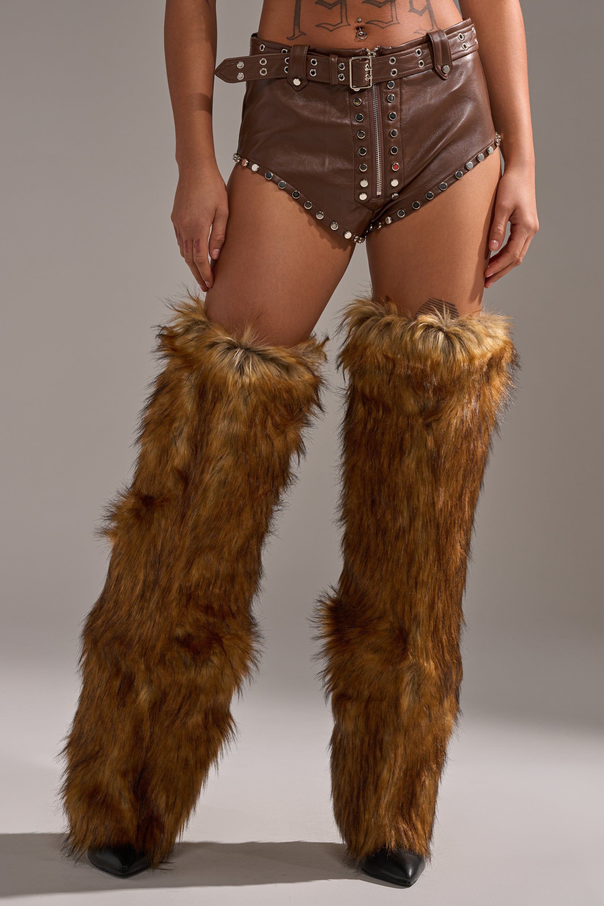A person wearing the BABY PLZ FAUX LEATHER STUD HOT SHORT IN BROWN, high-rise faux leather shorts with silver studs and a belt, paired with tall, furry brown leg warmers covering the legs from thigh to foot. 2