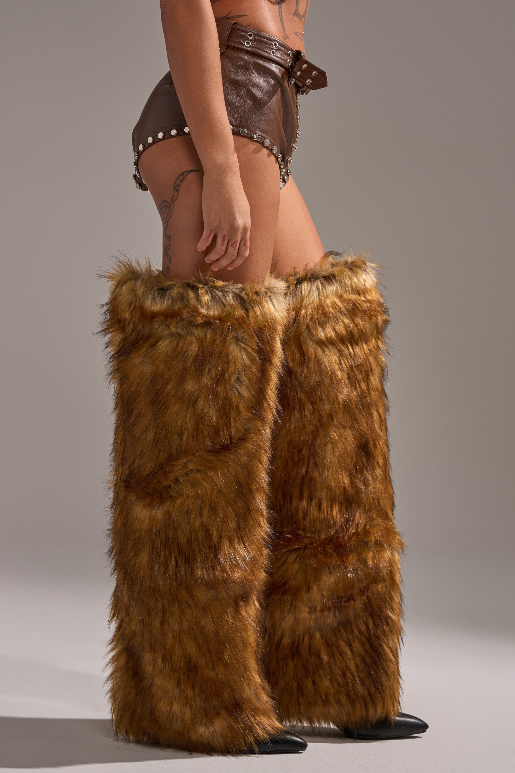 A person wears the BABY PLZ FAUX LEATHER STUD HOT SHORT IN BROWN with high, furry brown leg warmers covering the legs from feet to thighs, standing against a plain gray background. 3