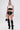 A woman stands confidently in a white crop top with black lace overlay, BABY PLZ FAUX LEATHER STUD HOT SHORTs, black thigh-high boots, statement jewelry, and holds a small black handbag with a chain strap.