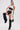 A woman poses confidently in a white crop top over a black lace bralette, BABY PLZ FAUX LEATHER STUD HOT SHORT, oversized black thigh-high boots, layered necklaces, and holding a small embellished handbag.