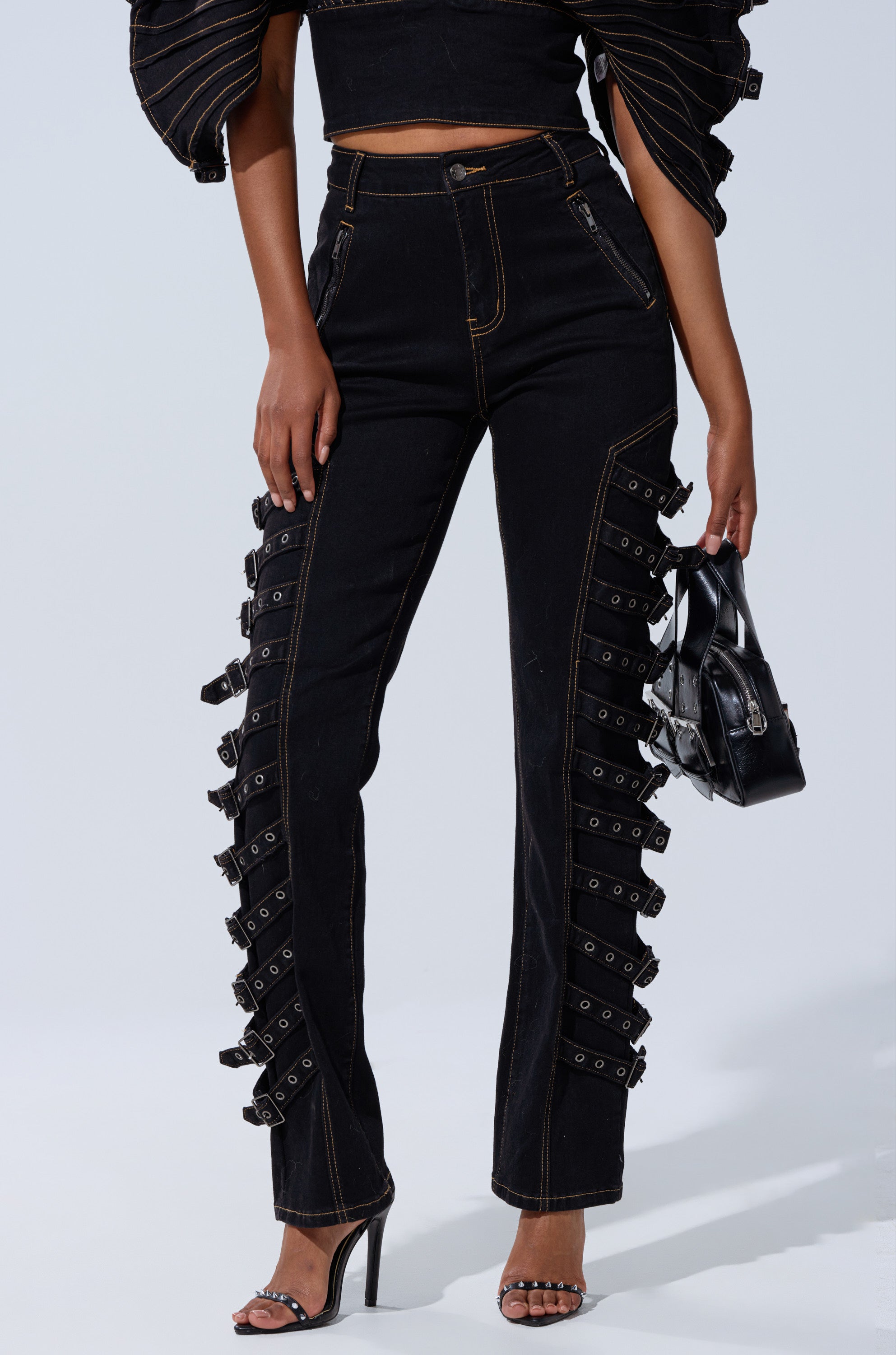 A woman wears the BUCKLE UP SKINNY DENIM PANT—high-rise black jeans with decorative side straps and adjustable buckle details—paired with a black crop top, black heeled sandals, and a small black handbag.