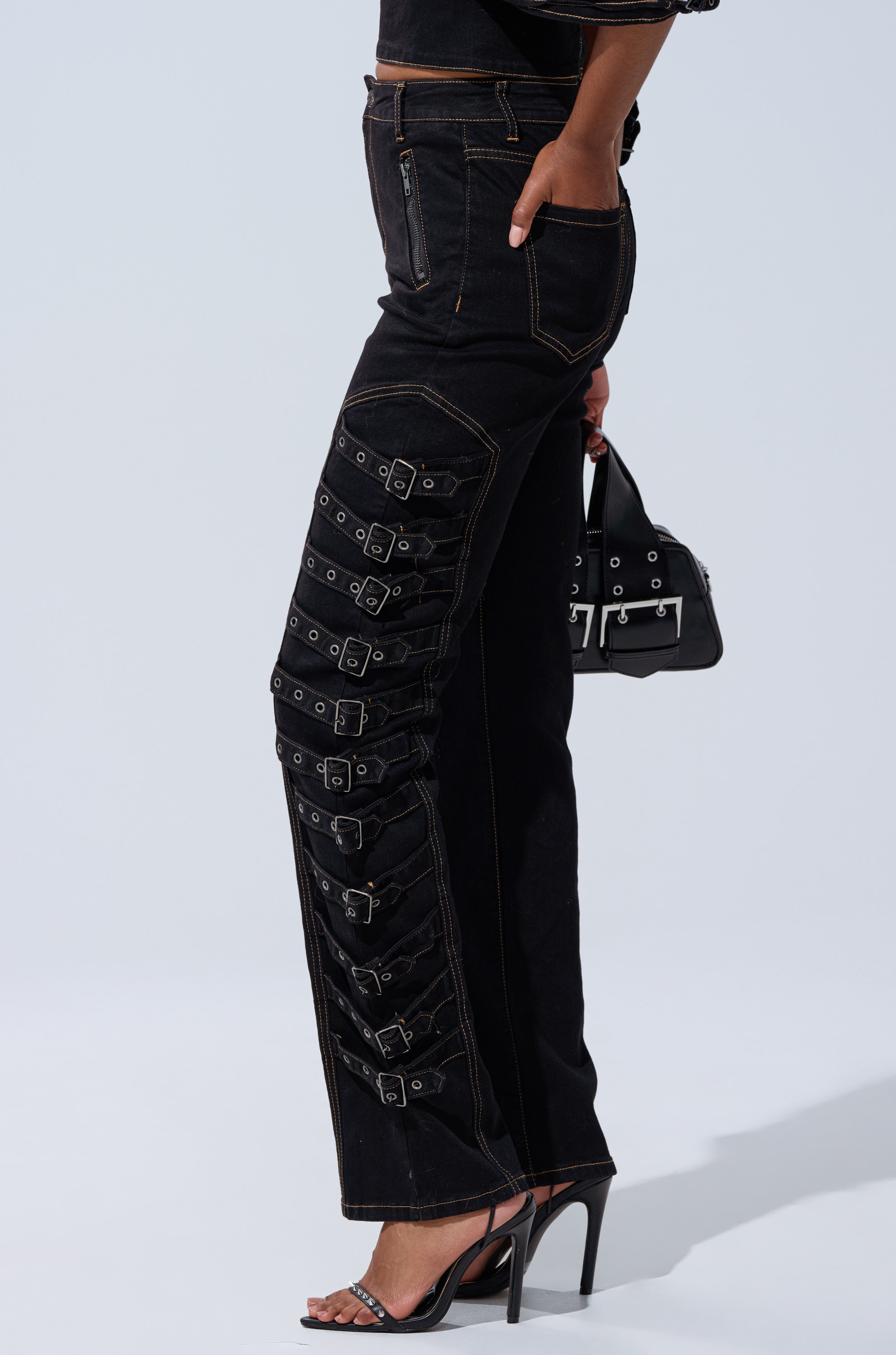 A person wearing the BUCKLE UP SKINNY DENIM PANT—high-rise black jeans with buckle embellishments along one leg—paired with a small black handbag and black high-heeled sandals.